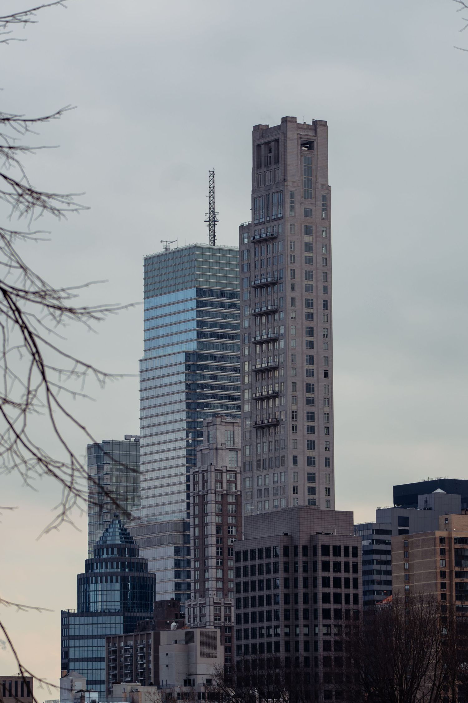 Modern and classic skyscrapers rise against a cloudy sky framed by bare winter tree branches.