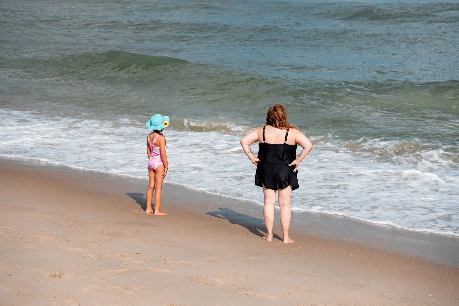 Woman in black swimsuit and young girl in pink swimsuit standing at ocean shoreline.