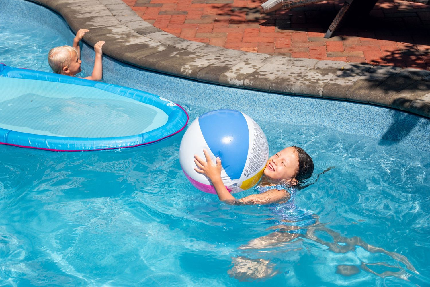 Girl hugging a colorful beach ball while swimming in a clear blue backyard pool.