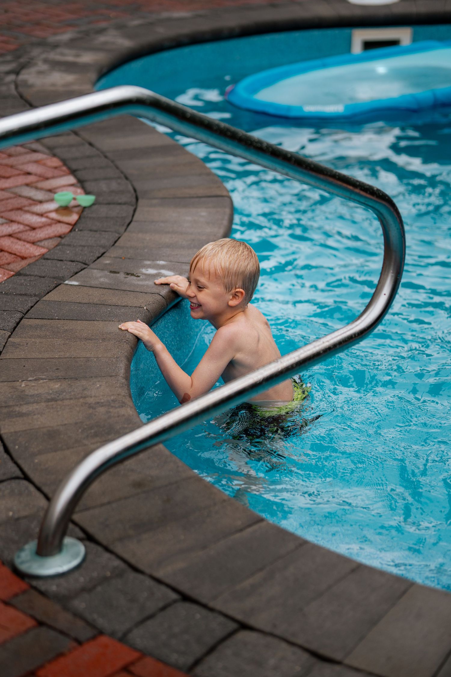 Young blonde boy smiling in a blue outdoor swimming pool, holding onto metal handrails near the pool steps.
