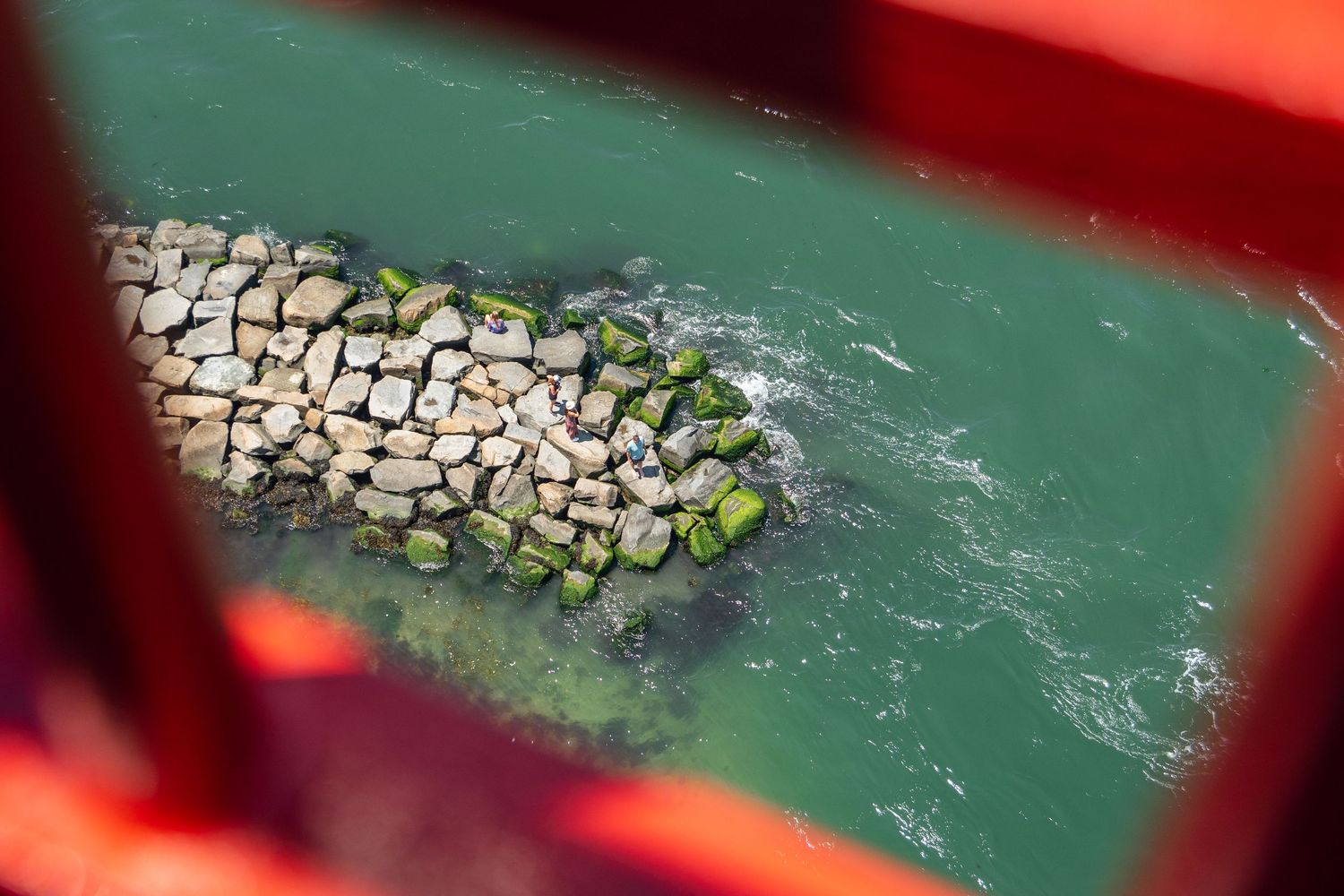 Rocky breakwater with green moss viewed through red metal bridge beams over turquoise water.
