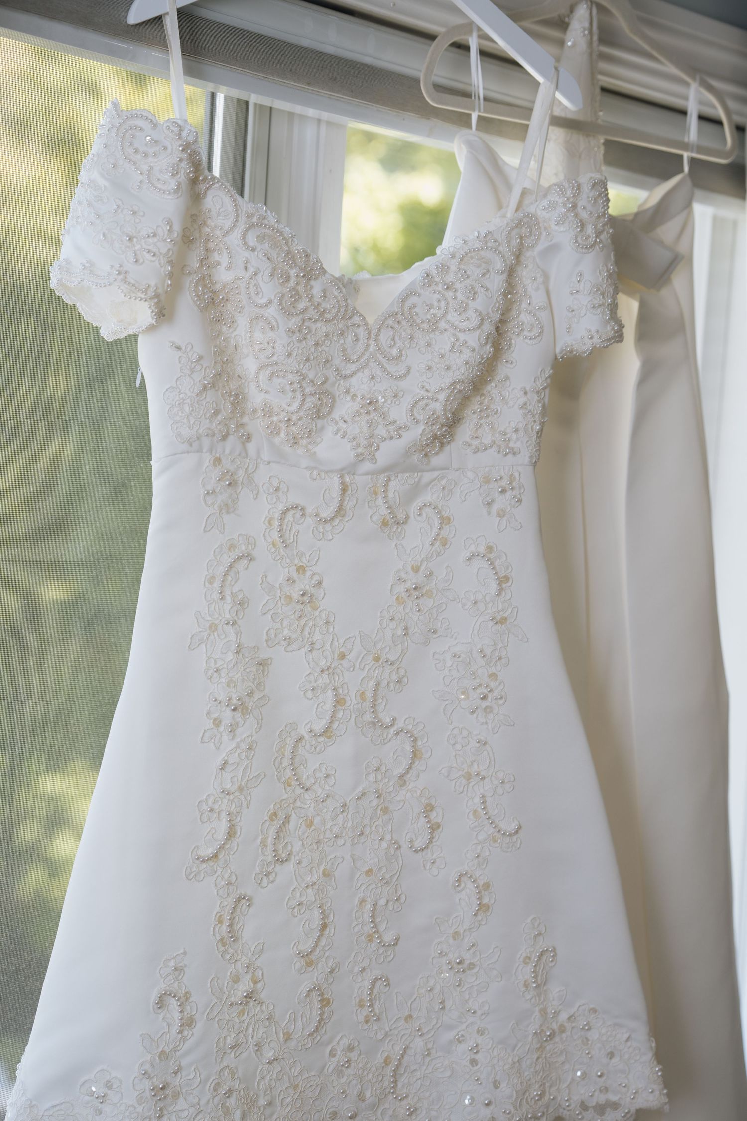 White lace wedding dress with beaded embroidery and spaghetti straps hanging on a hanger near a window.