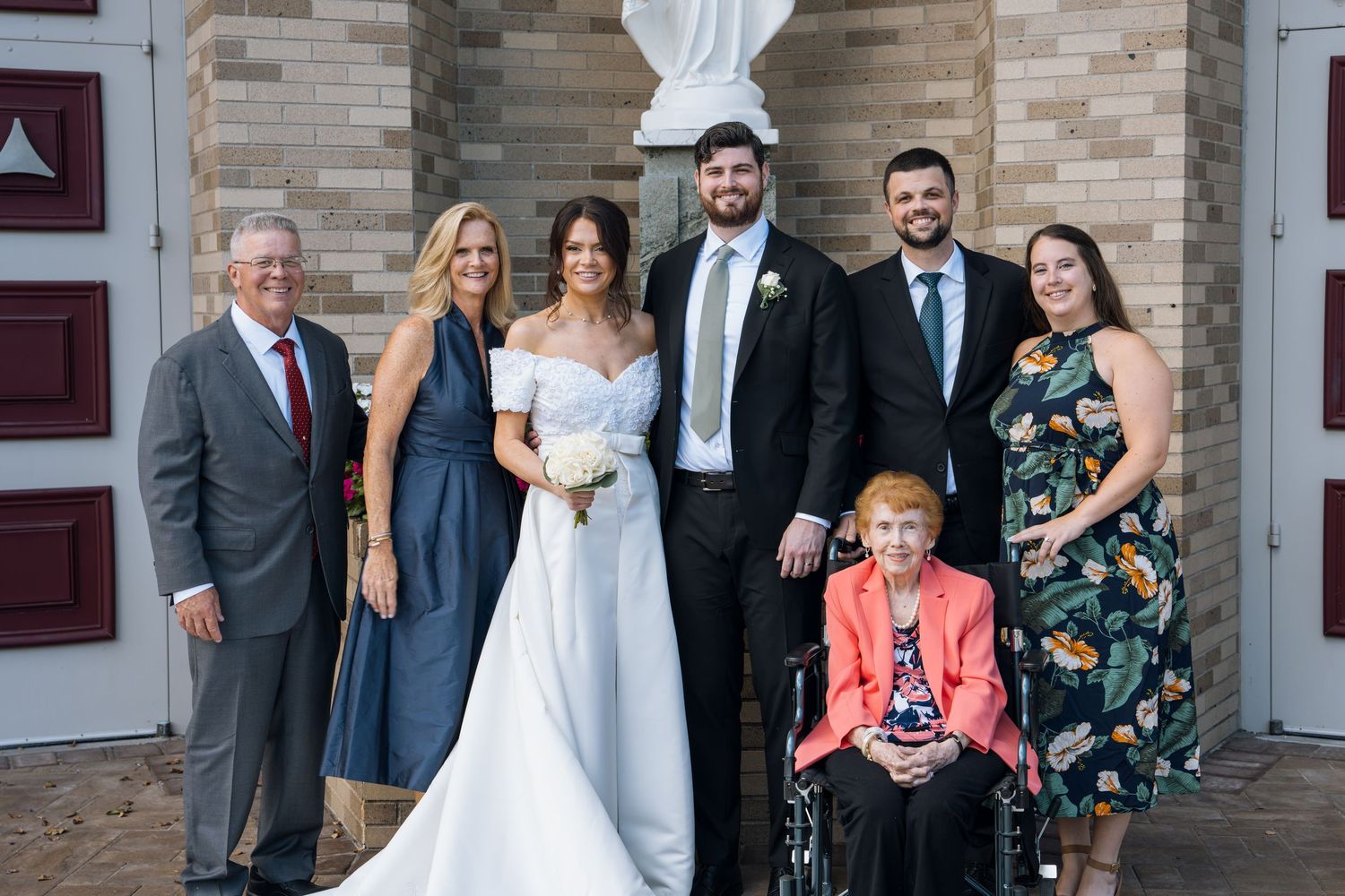 Wedding party poses outside a church with bride in white gown, groom in suit, family members, and elderly woman in wheelchair.