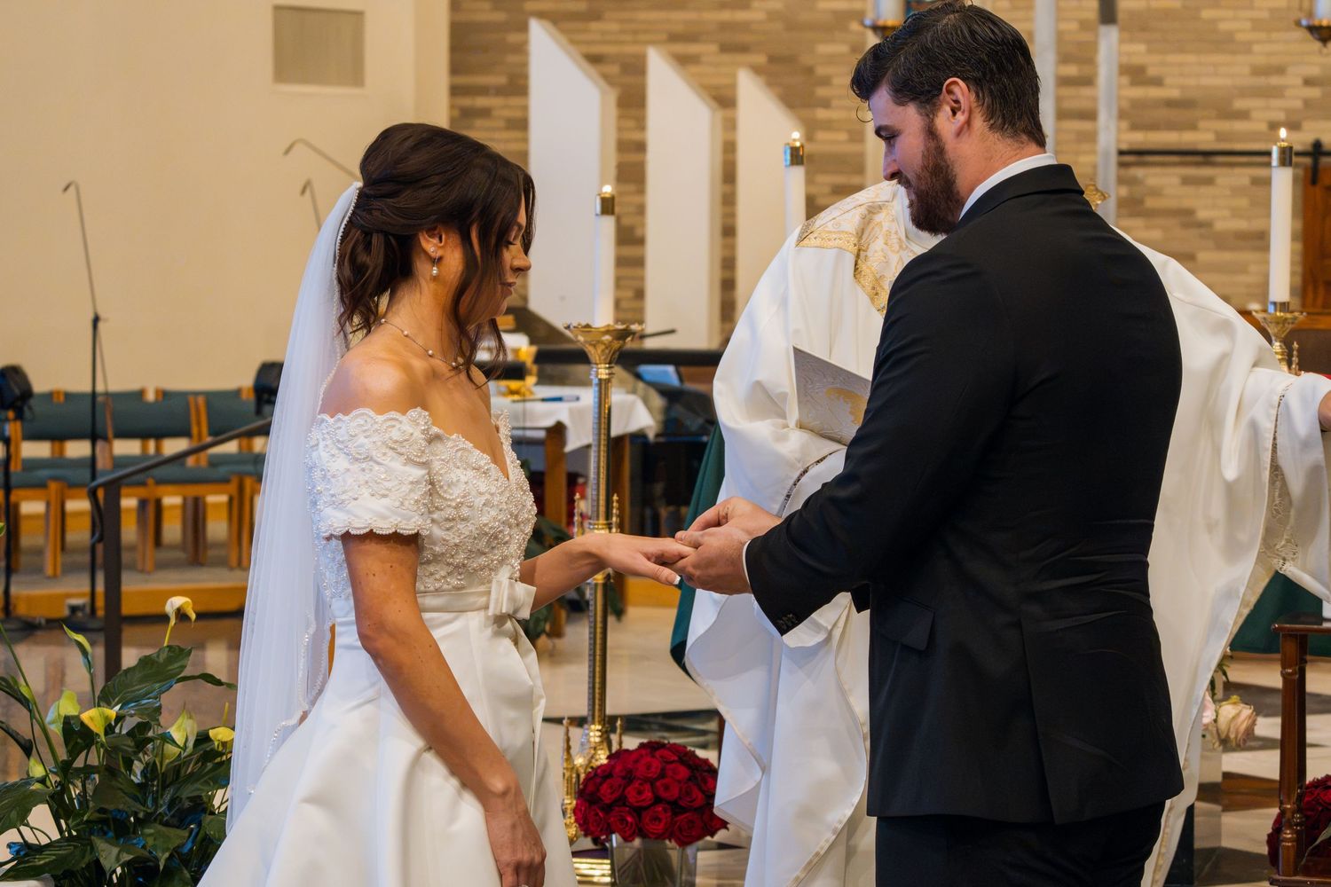 Bride and groom exchange rings at church altar during wedding ceremony, with officiant in white robes standing behind them.