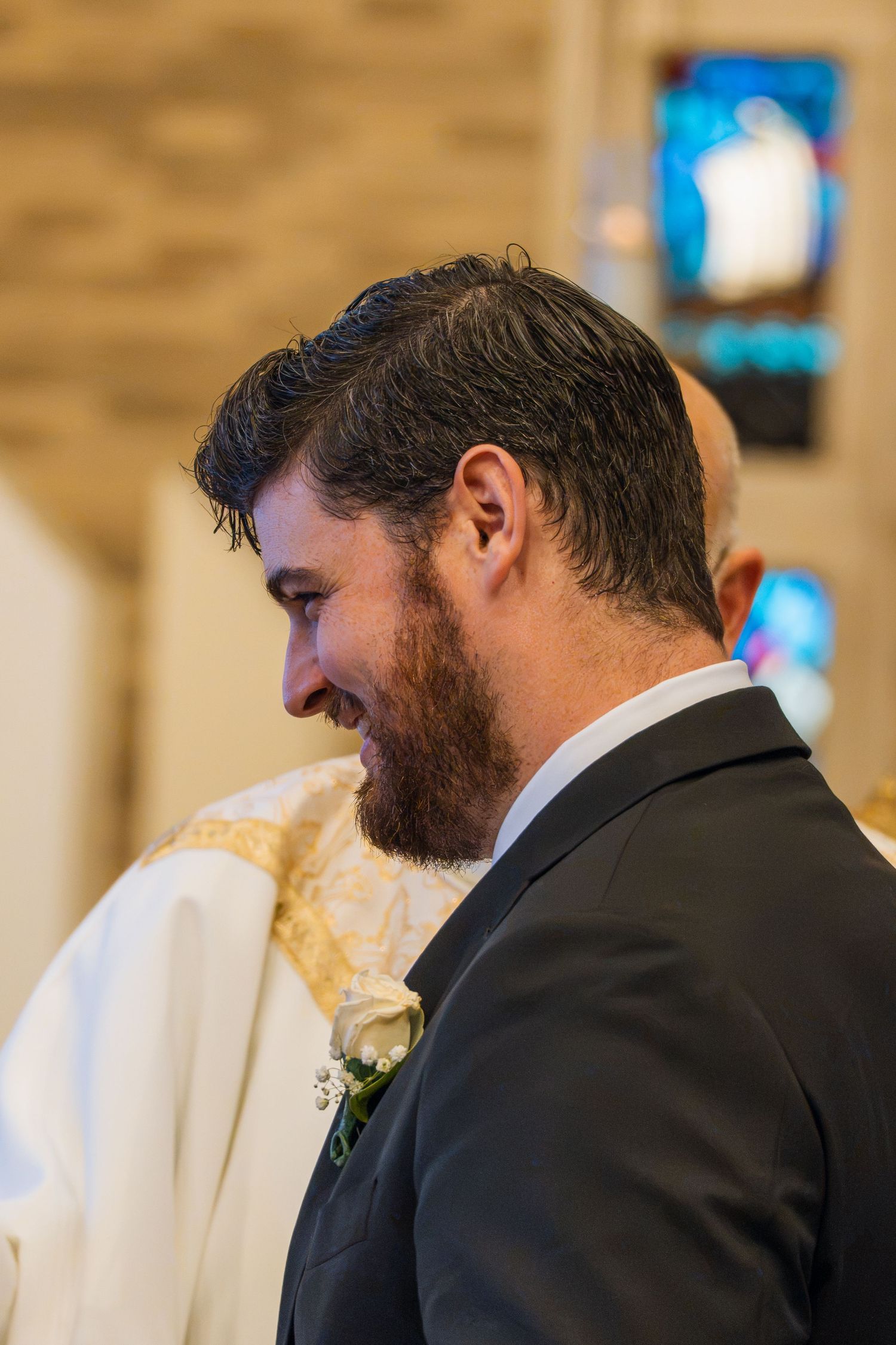 Smiling bearded groom in dark suit with boutonniere at church wedding ceremony, stained glass visible in background.