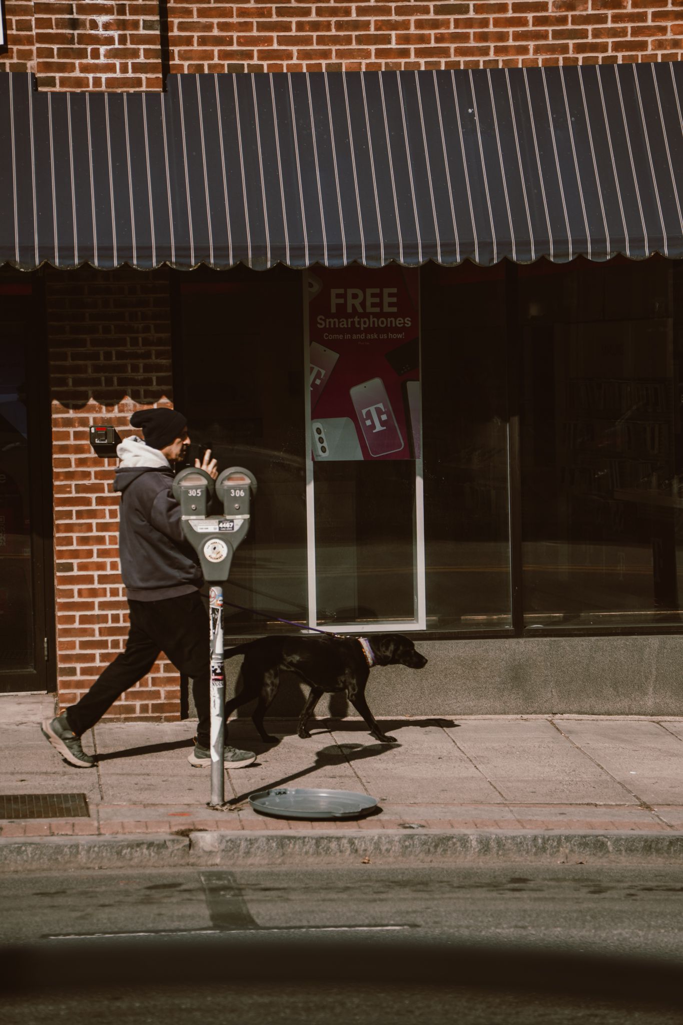 Person walking a black dog past a parking meter outside a brick storefront.