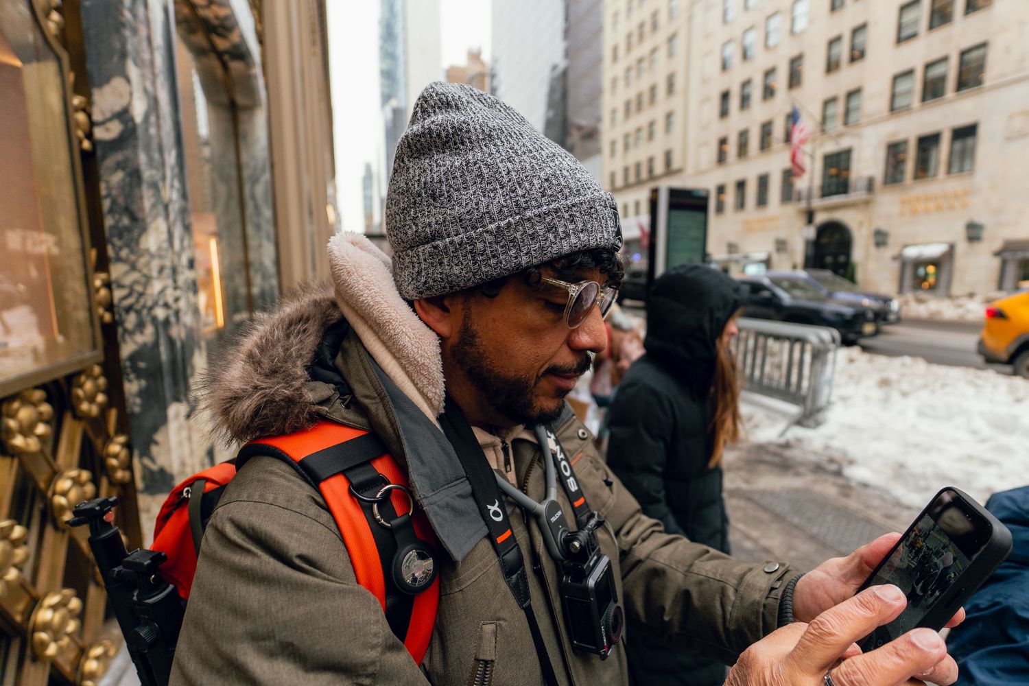 Man in gray beanie and orange backpack photographing with phone on snowy NYC street.