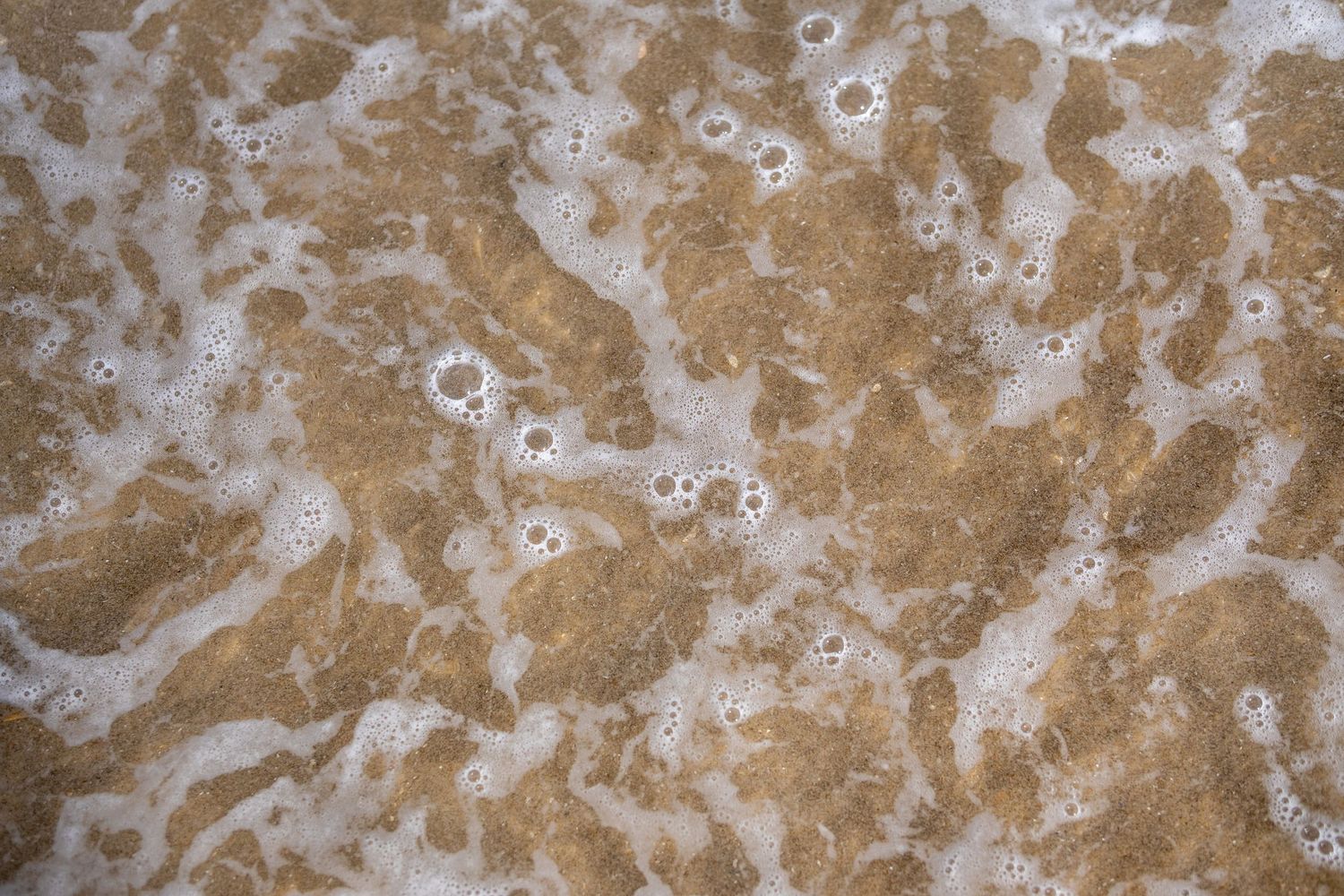 Aerial view of shallow foamy ocean water over sandy brown beach bottom texture.