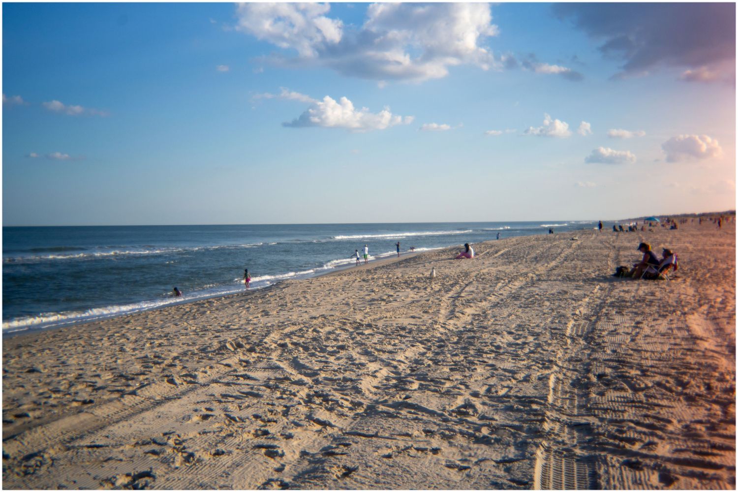 Wide sandy beach with gentle waves, scattered sunbathers, and a bright blue sky with clouds.