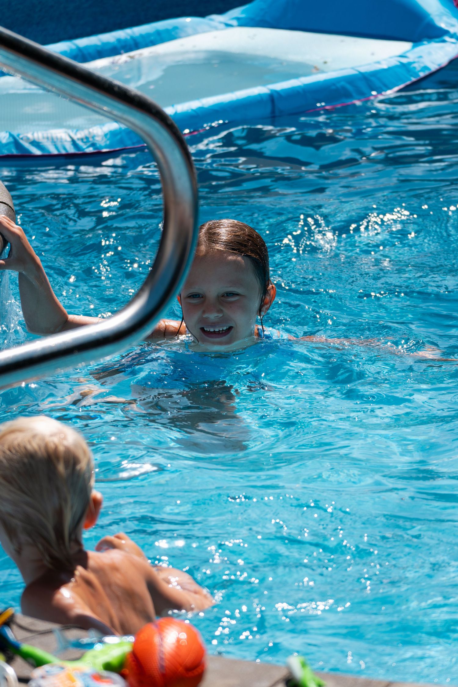 Happy girl swimming in a sunny blue pool with a young child and red ball at the edge.