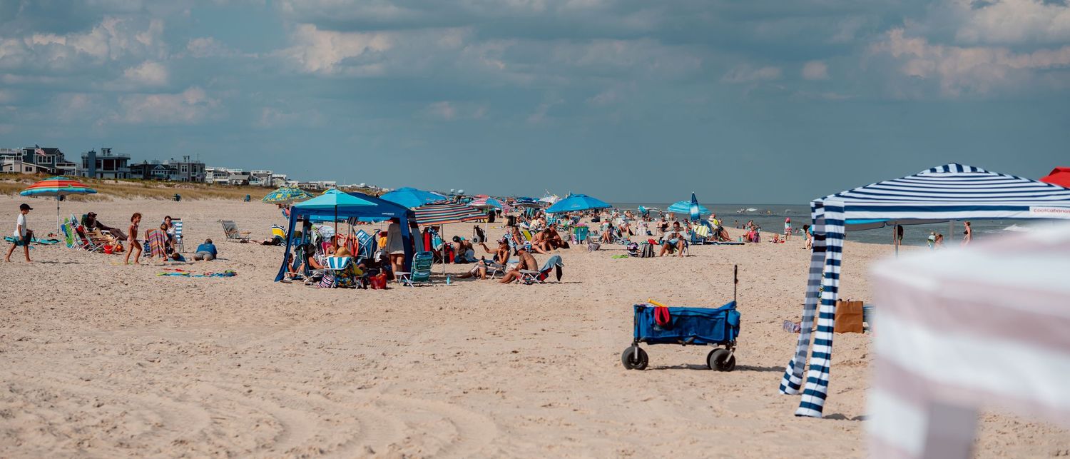 Crowded sandy beach with colorful umbrellas, sunbathers, and a blue wagon on a partly cloudy summer day.