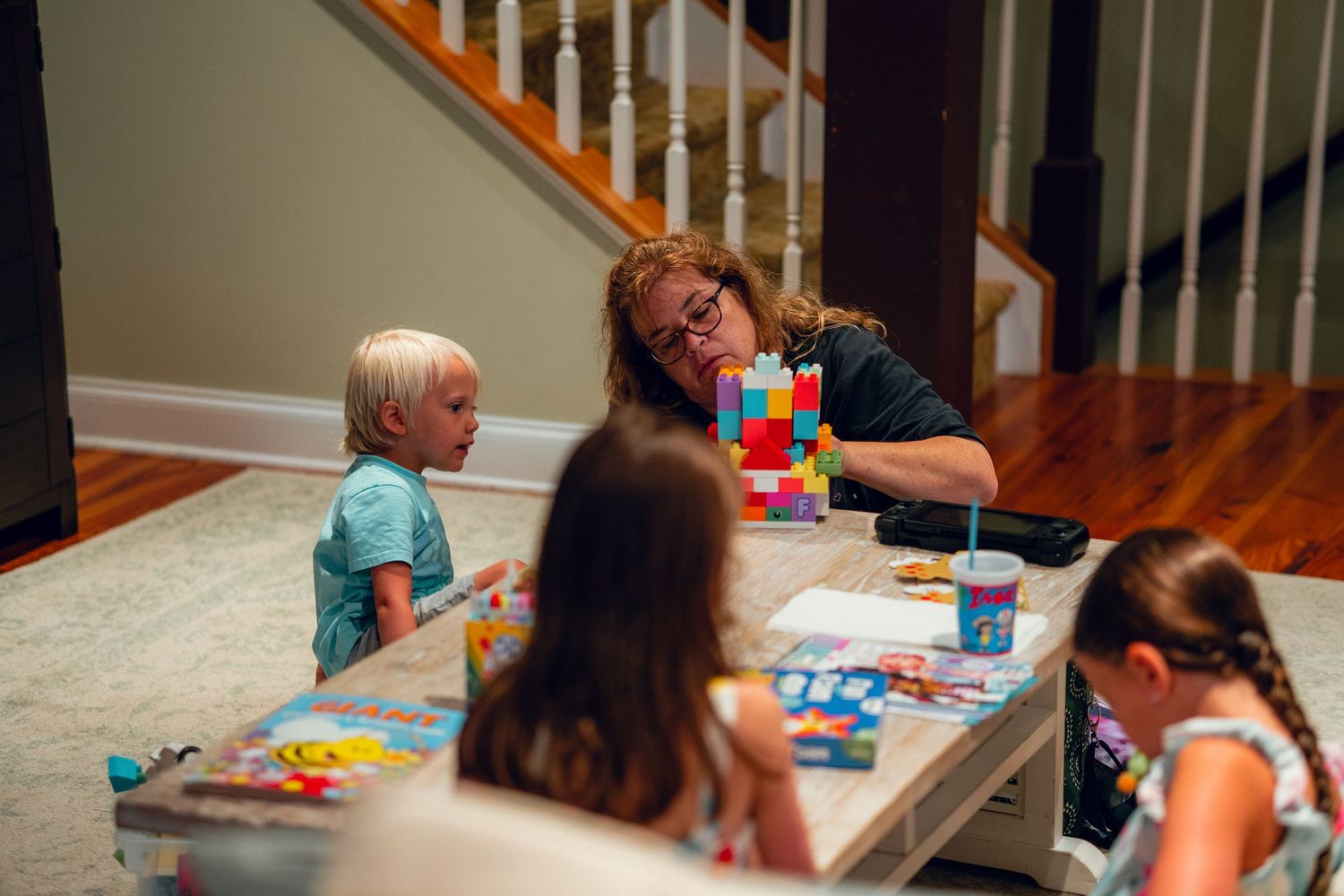 Woman playing with colorful building blocks with three young children at a low table indoors.