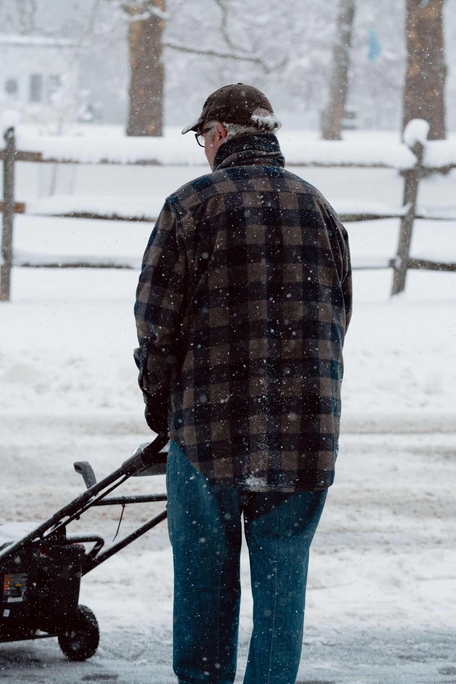 Elderly man in plaid jacket operating a snowblower from behind during a snowy winter day.