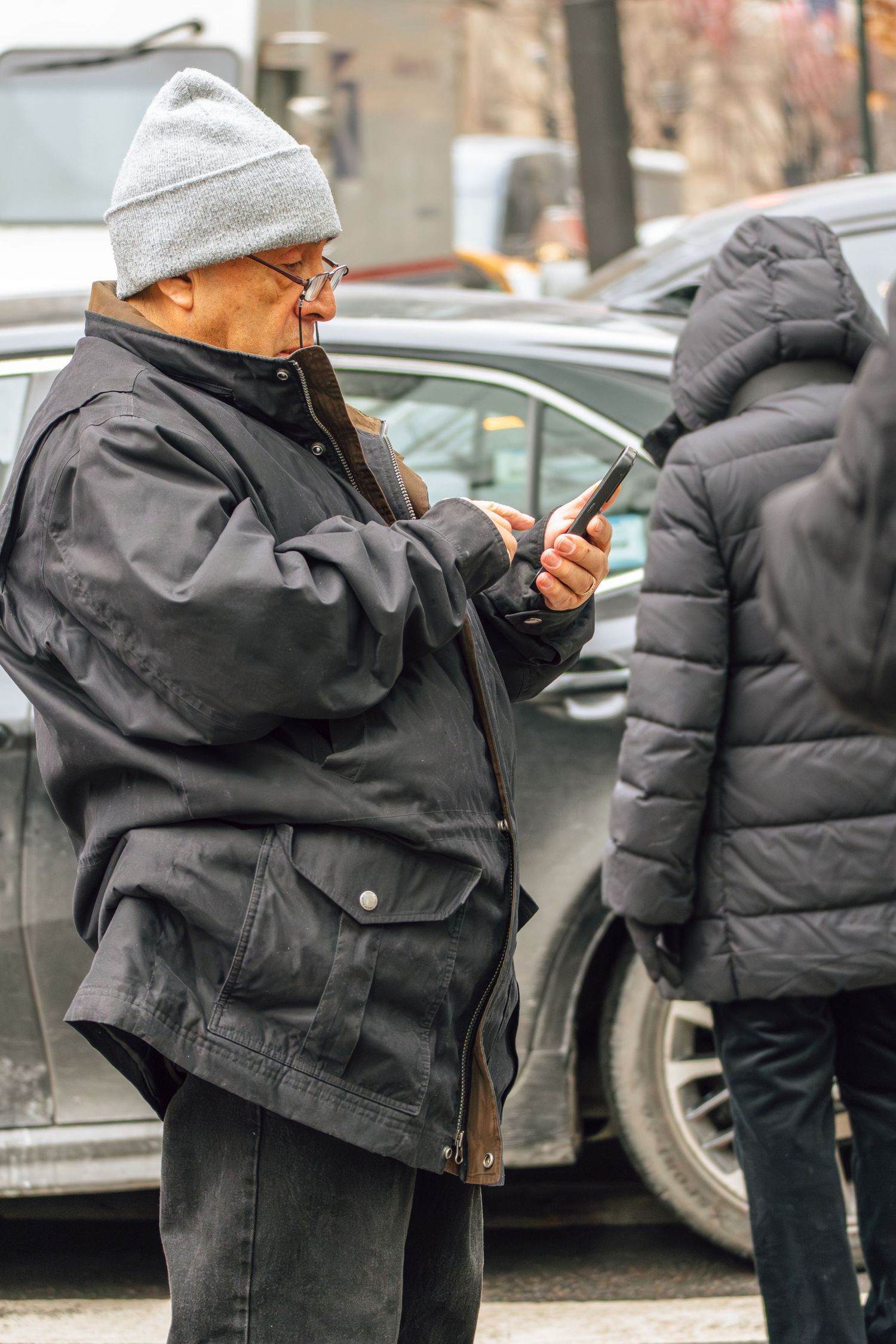 Elderly man in gray beanie and black jacket checking his smartphone while standing on a New York City street.