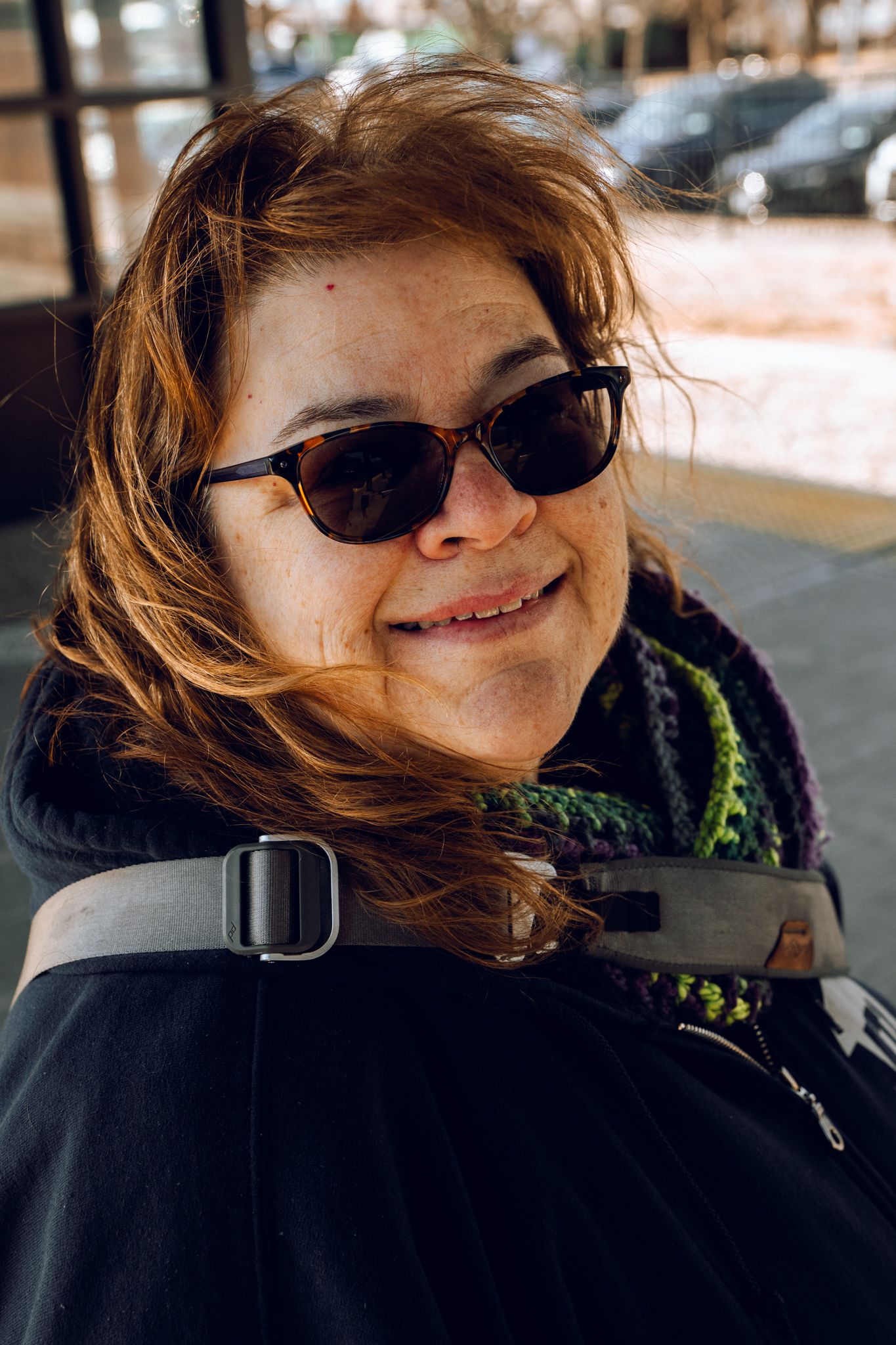 Smiling red-haired woman wearing sunglasses and a dark coat with a green scarf on a sunny day.
