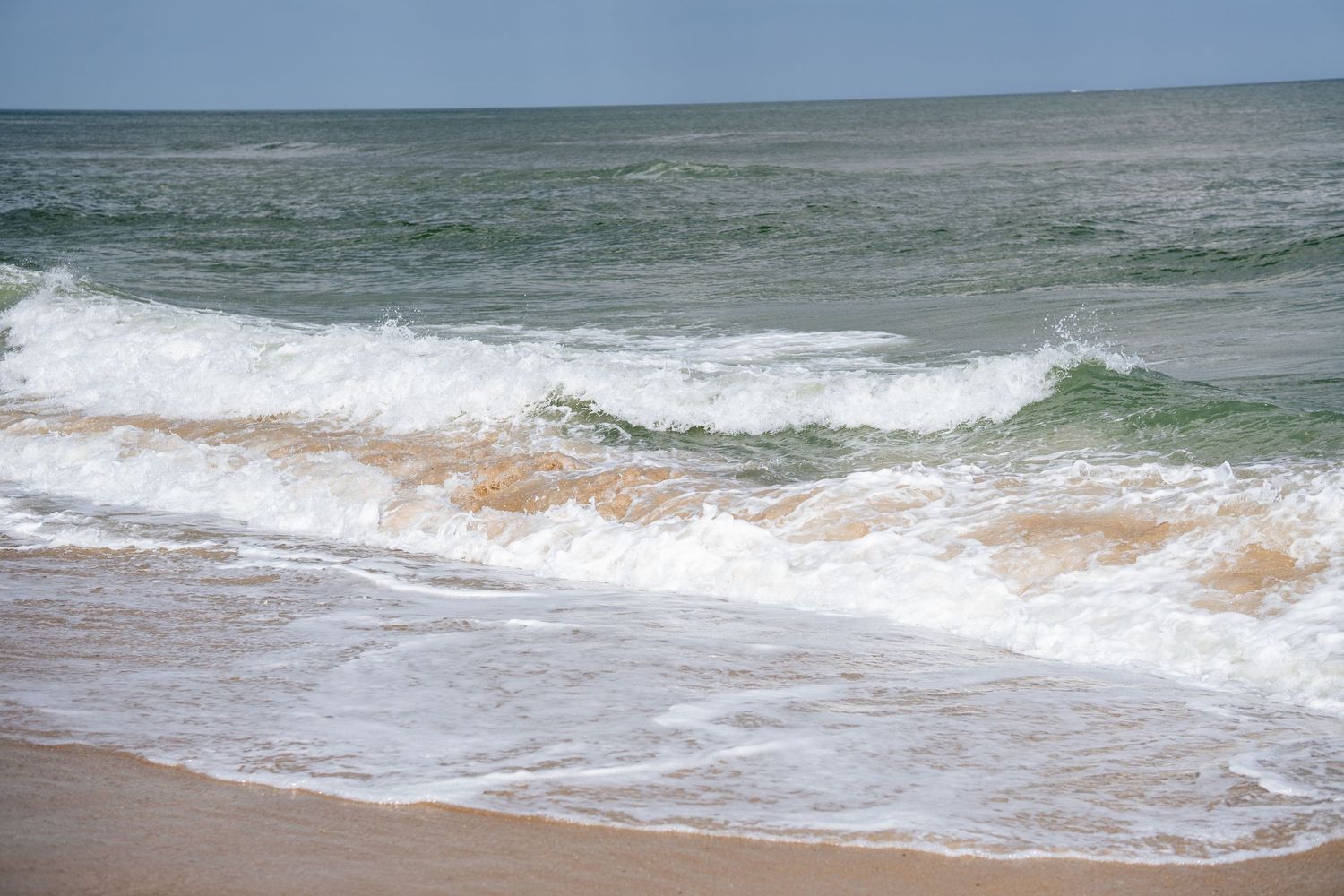 Rolling ocean waves with white foam breaking onto a sandy beach under a hazy sky.