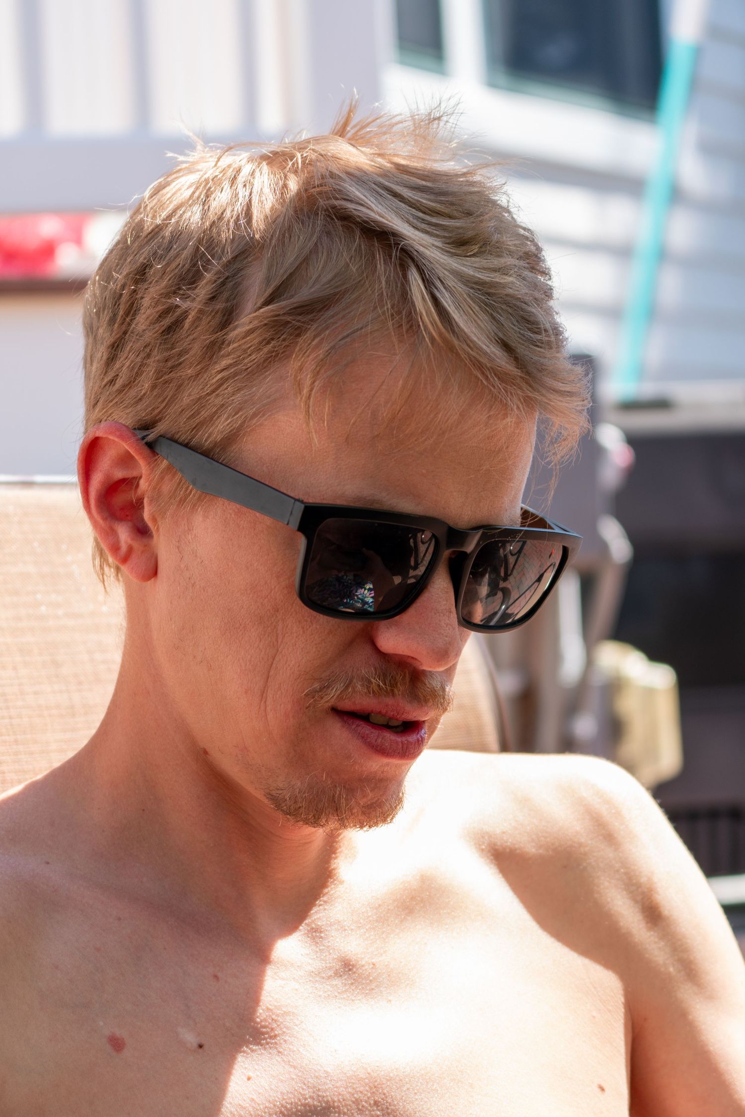 Blonde man wearing black sunglasses relaxing shirtless outdoors on a sunny day.