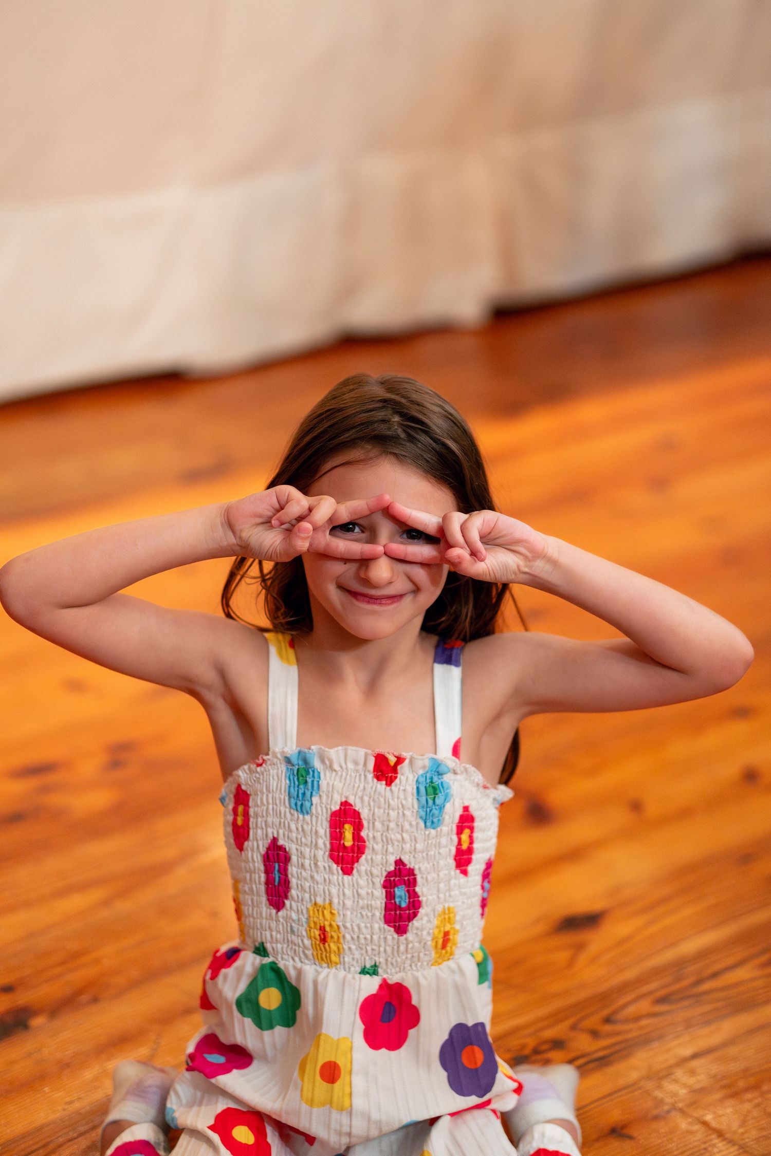 Young girl in a colorful floral dress making peace signs over her eyes while sitting on a wood floor.