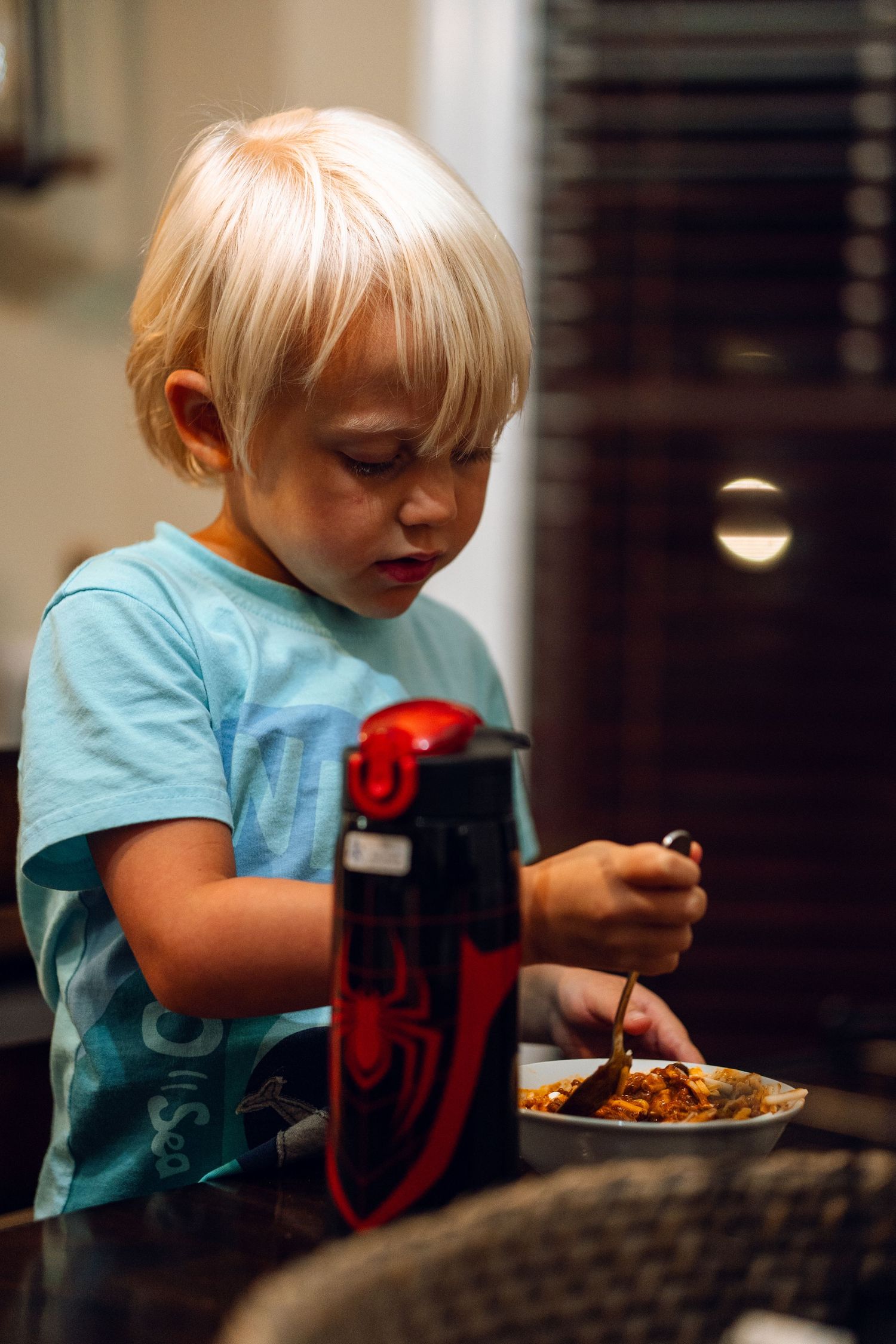 A young blonde boy in a blue t-shirt eats a meal at a table with a red water bottle nearby.