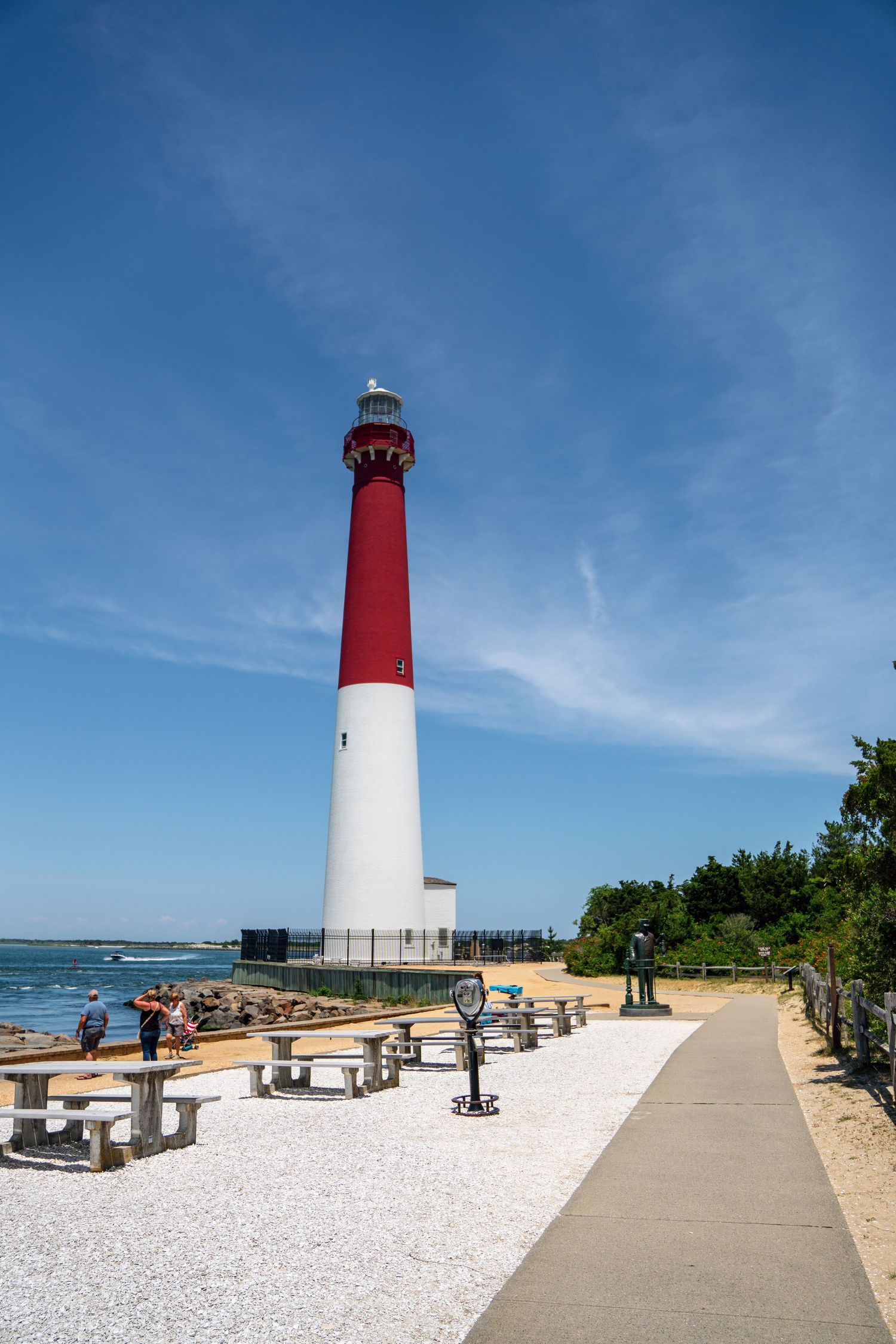 Red and white Barnegat Lighthouse stands tall against blue sky, with picnic area and sandy beach in New Jersey.