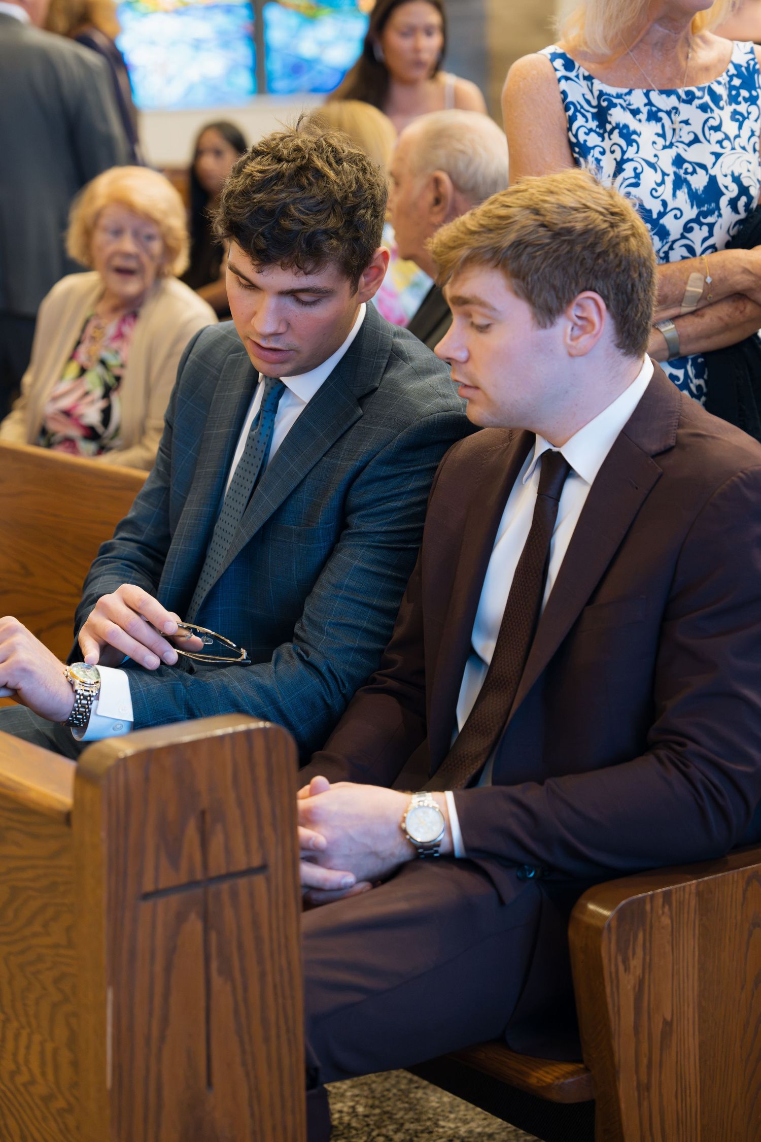 Two young men in suits sitting in church pews during a formal ceremony or wedding service.