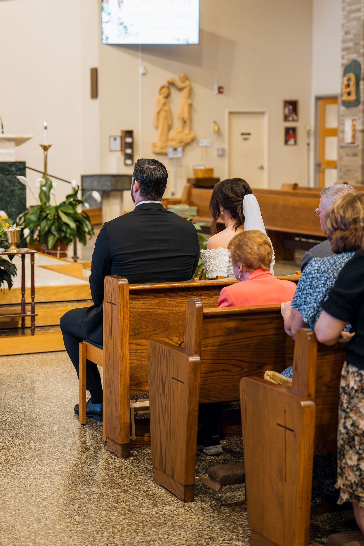 Bride and groom seated in wooden church pews during a wedding ceremony with guests and altar visible.
