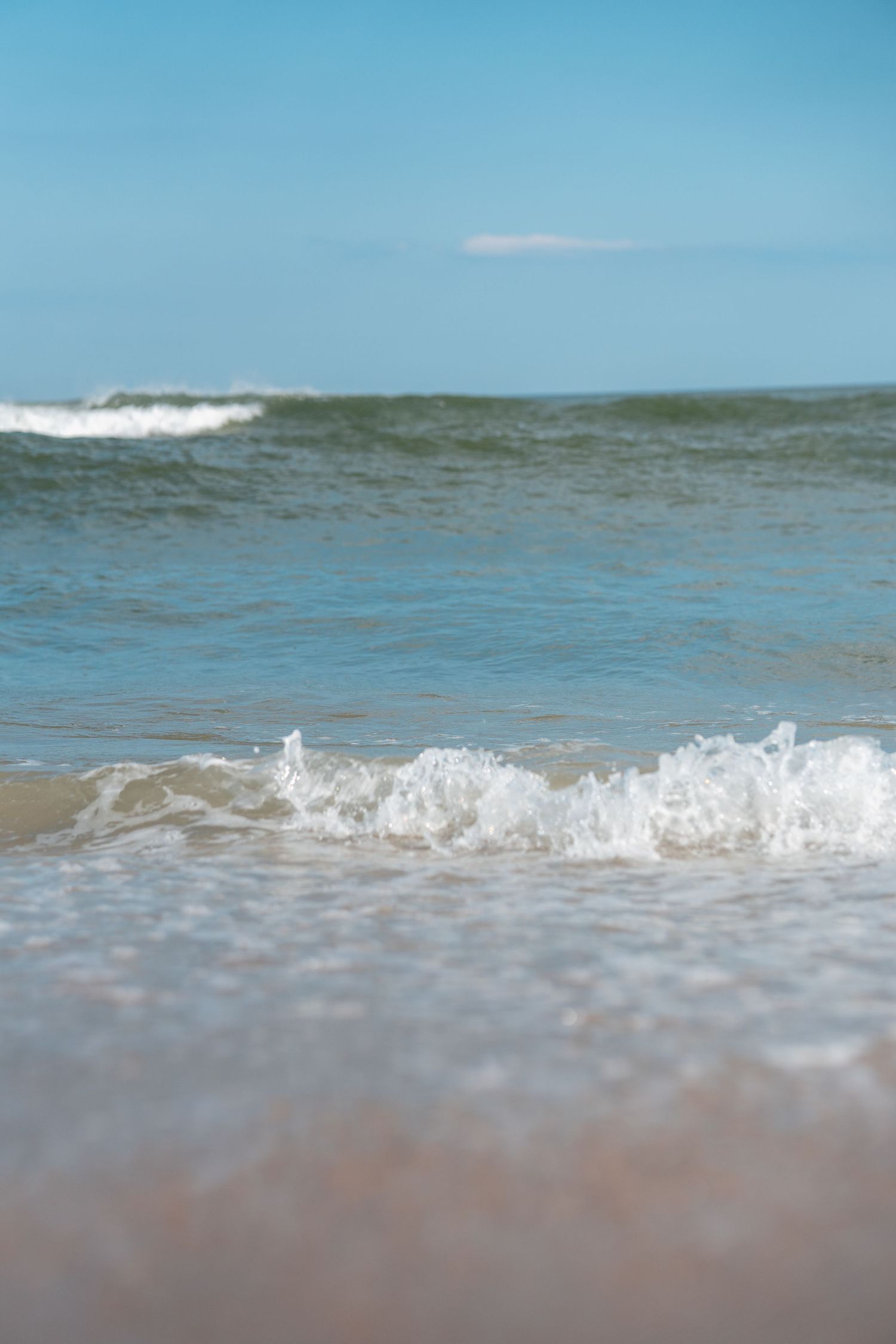 Gentle ocean waves washing onto a sandy beach with blue sky and horizon in the background.