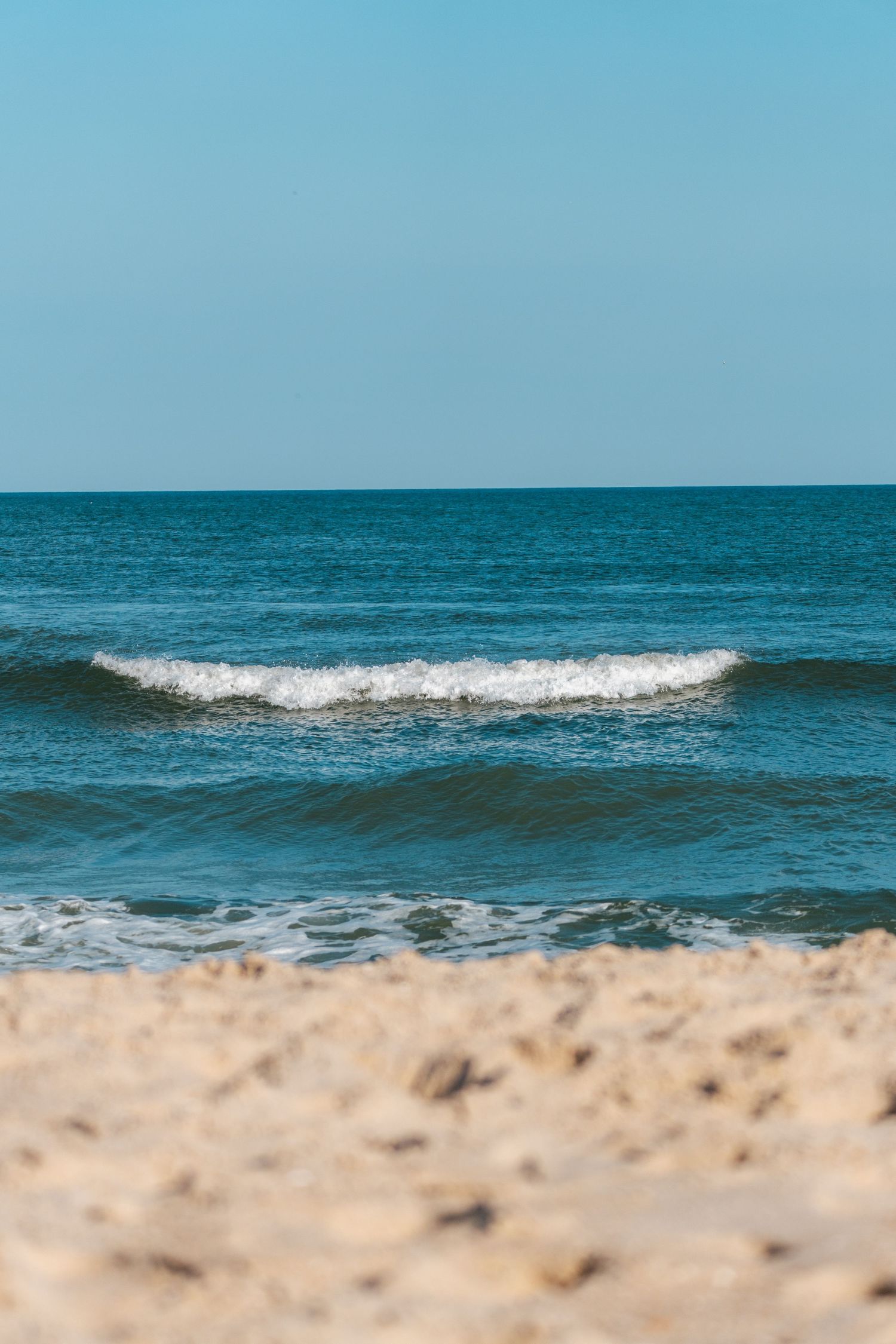 Gentle ocean wave rolling onto a sandy beach under a bright clear blue sky on a sunny day.