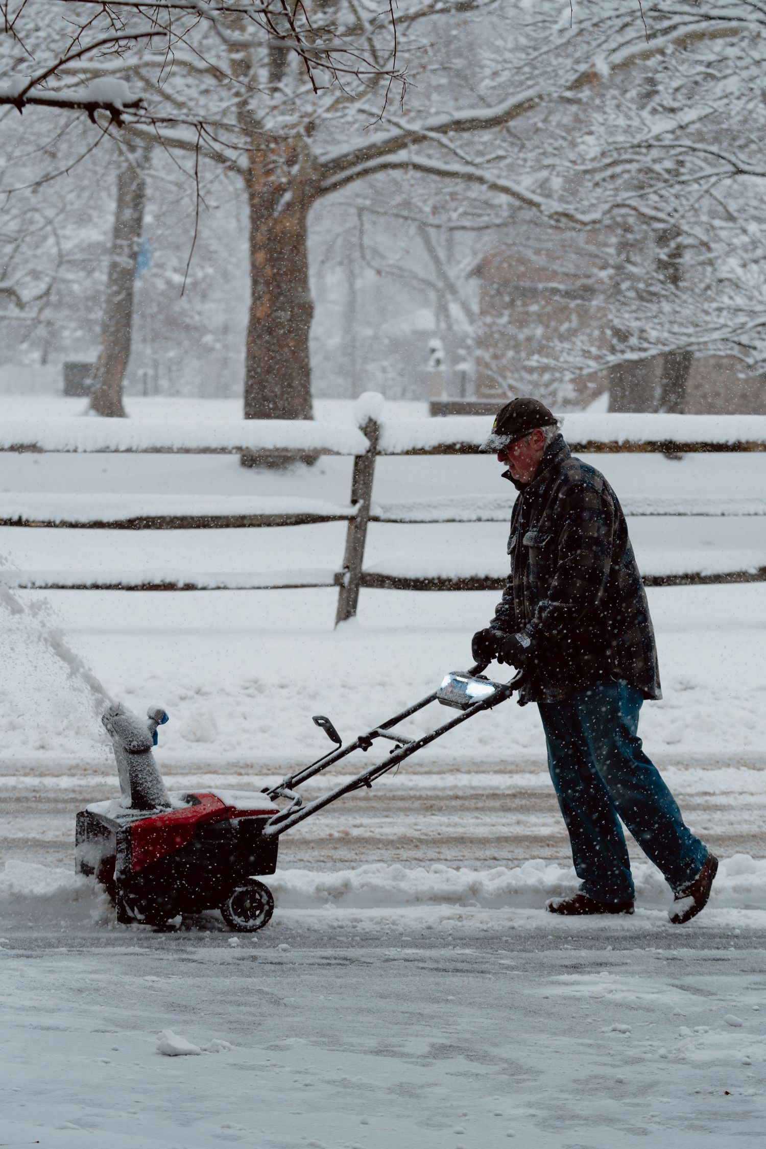A man using a red snowblower to clear snow from a driveway surrounded by snow-covered trees.