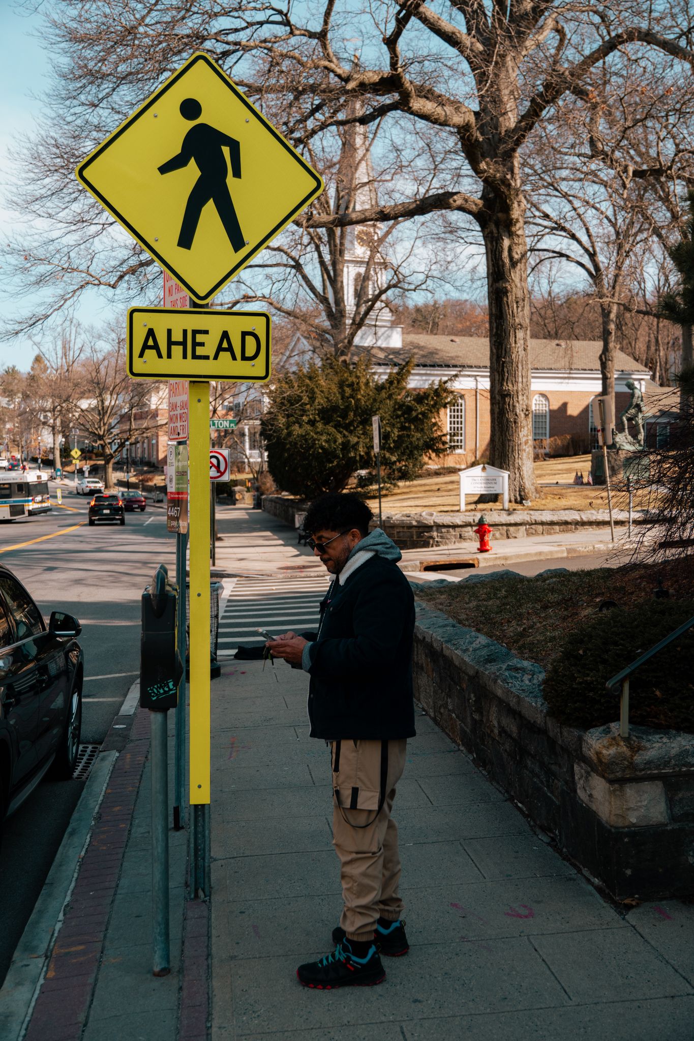 Man standing under a pedestrian crossing ahead sign on a sunny urban sidewalk.