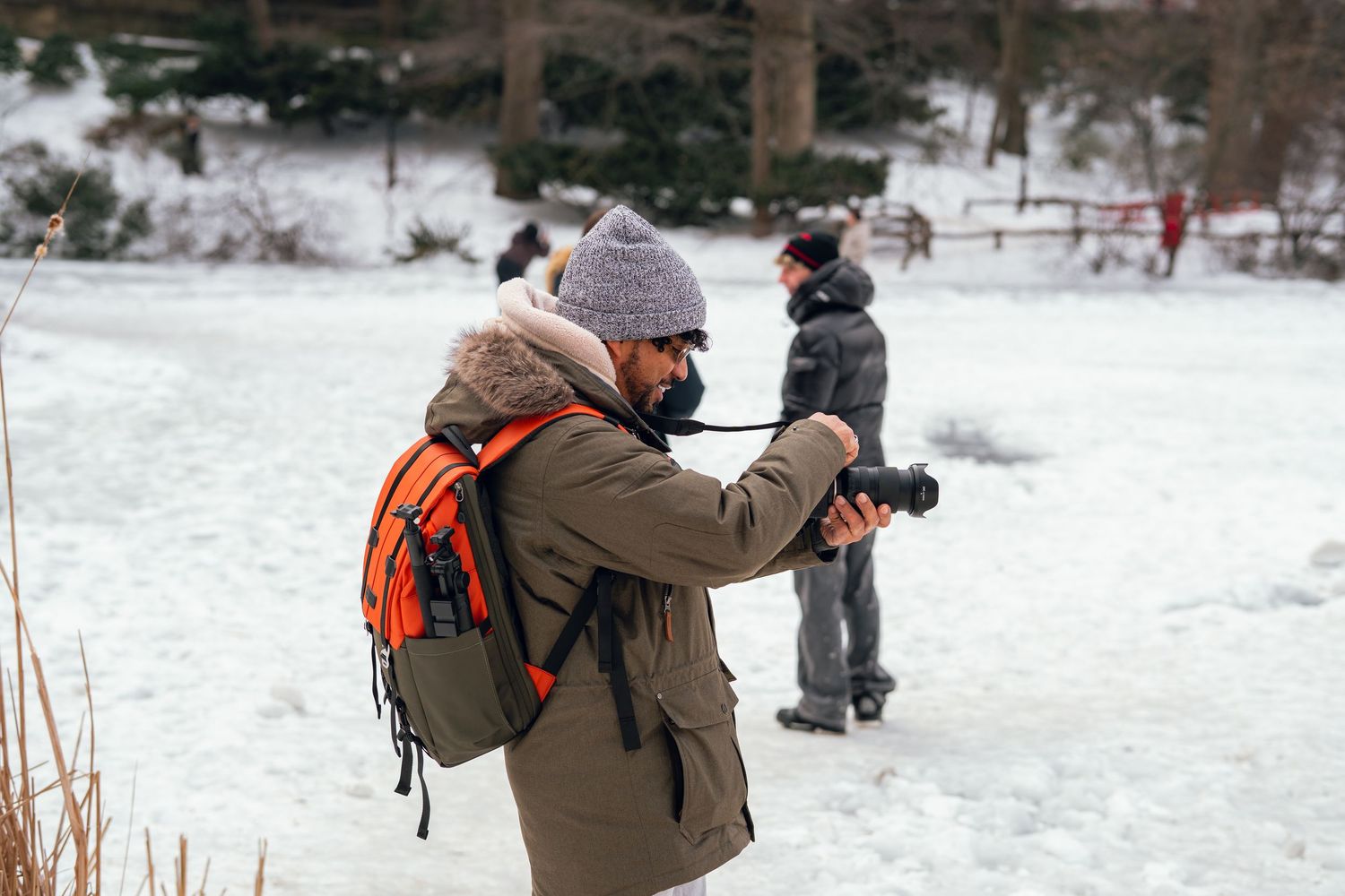 Male photographer in gray beanie and olive jacket taking photos in a snowy winter landscape.