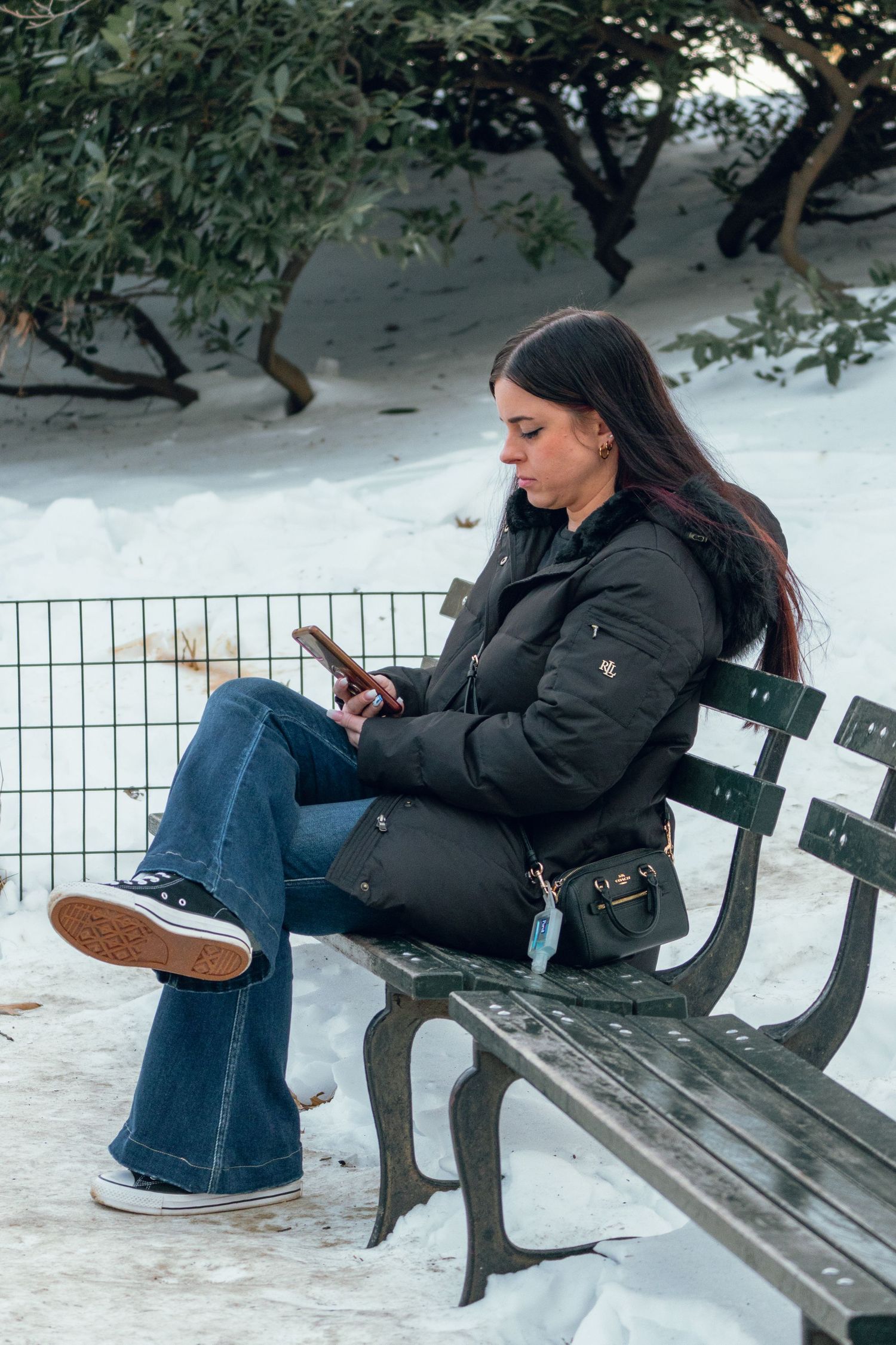 Young woman in black jacket and flare jeans sits on park bench using phone in snow.