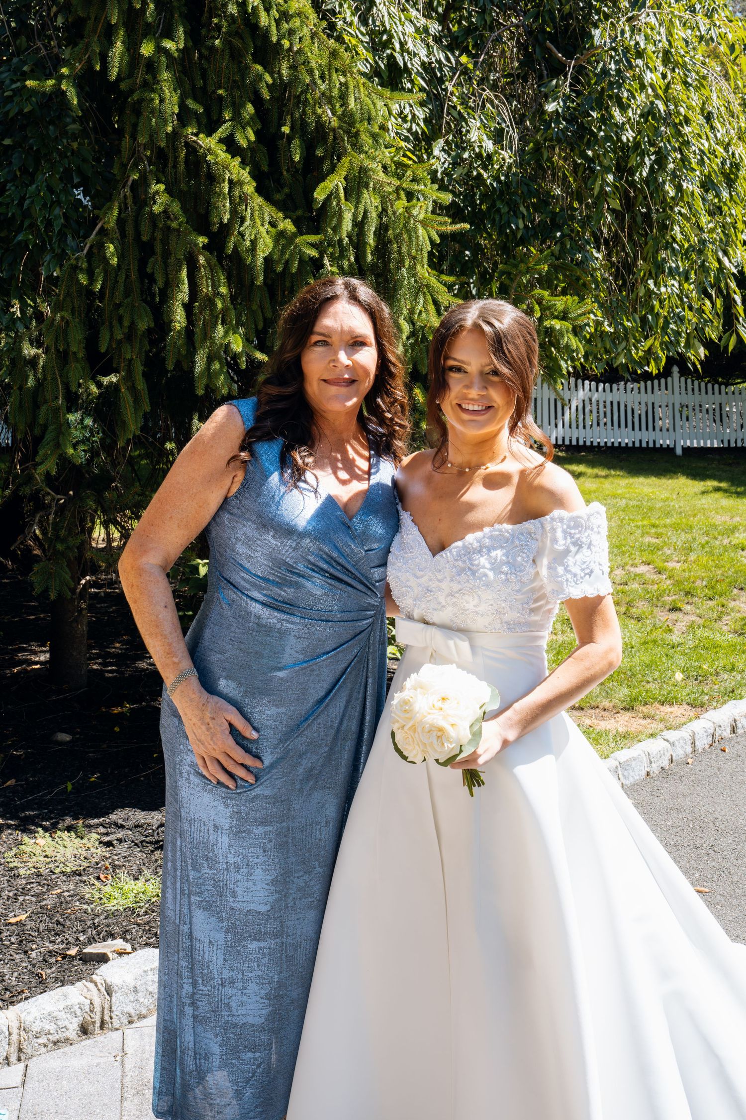 Bride in white lace gown holding bouquet poses with woman in blue metallic dress outdoors on sunny day.