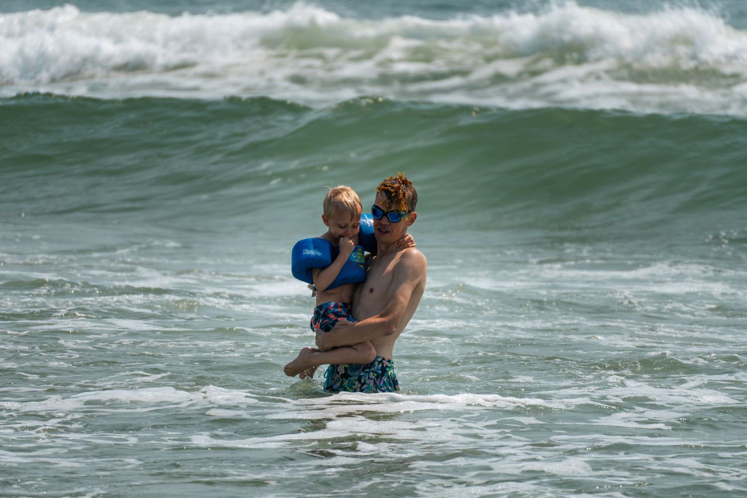 Adult holding a young child in ocean waves at a sunny beach, both enjoying the water together.