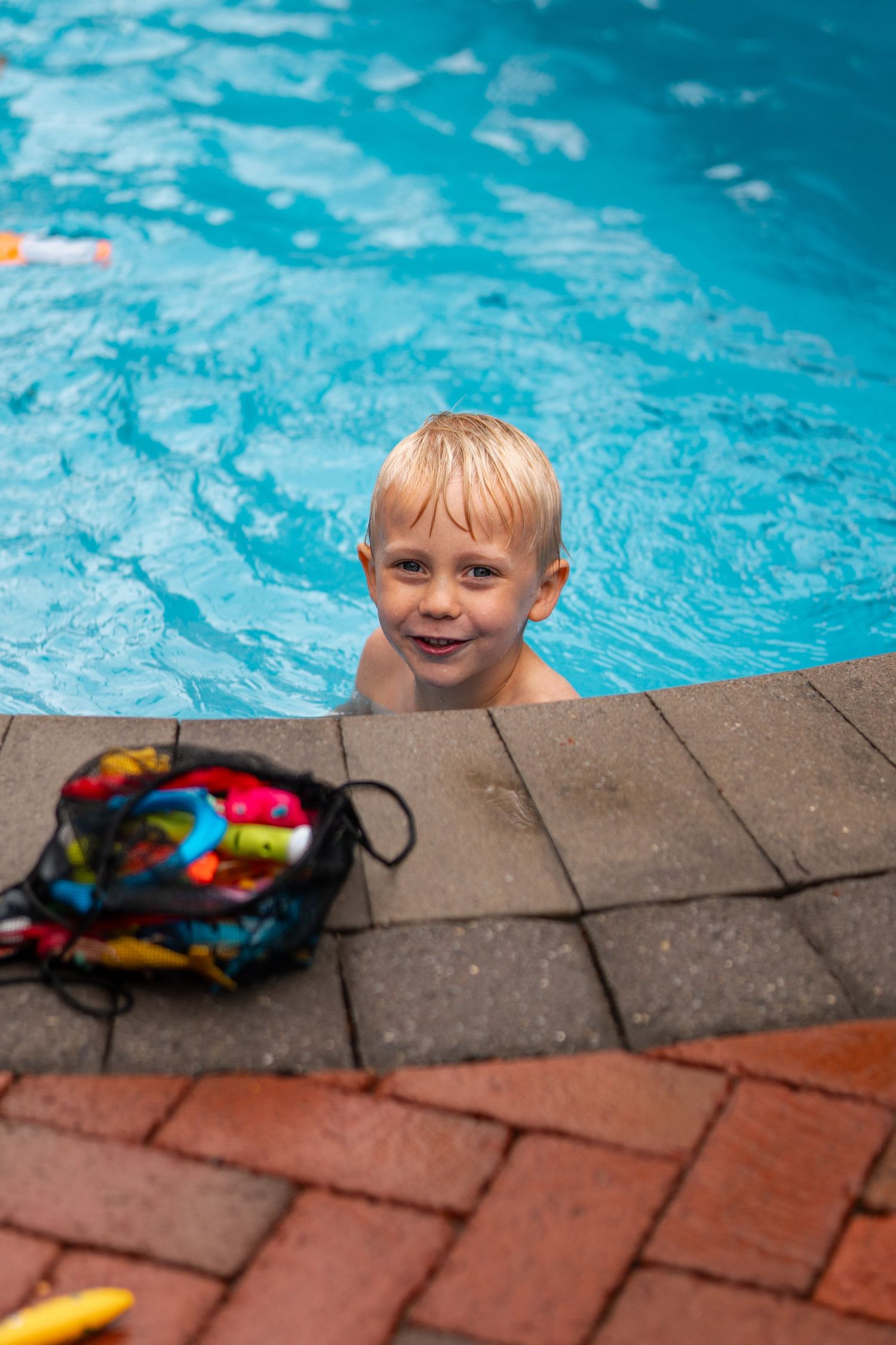 Smiling blonde boy peeks over pool edge at brick poolside, with colorful swim bag nearby on sunny day.