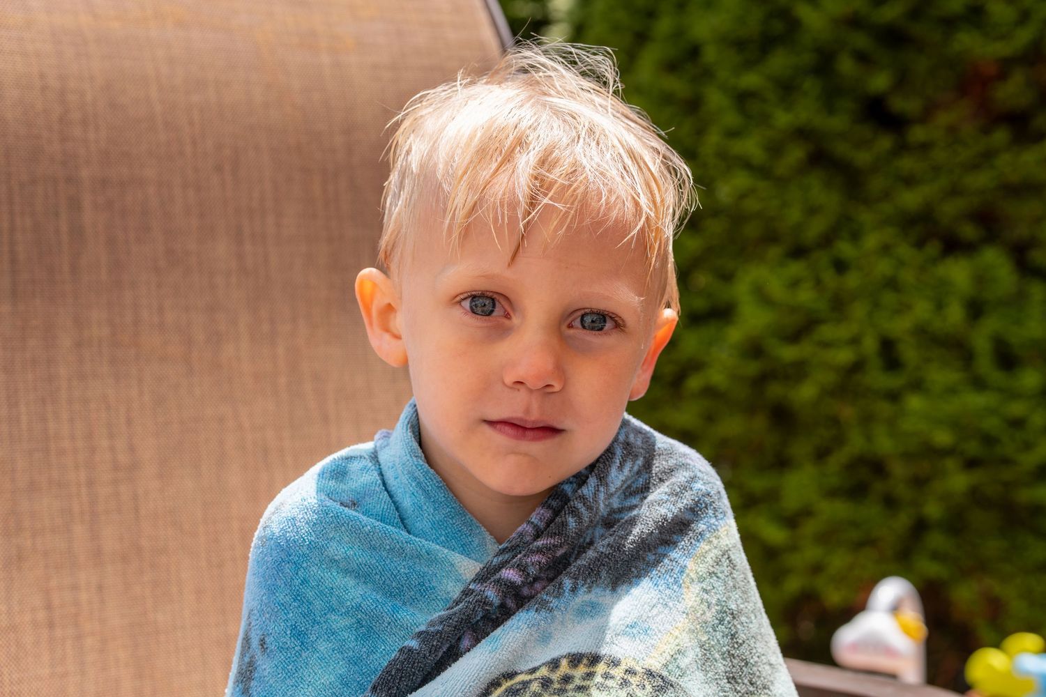 Young blond boy wrapped in a blue towel sitting on a patio chair outdoors on a sunny day.