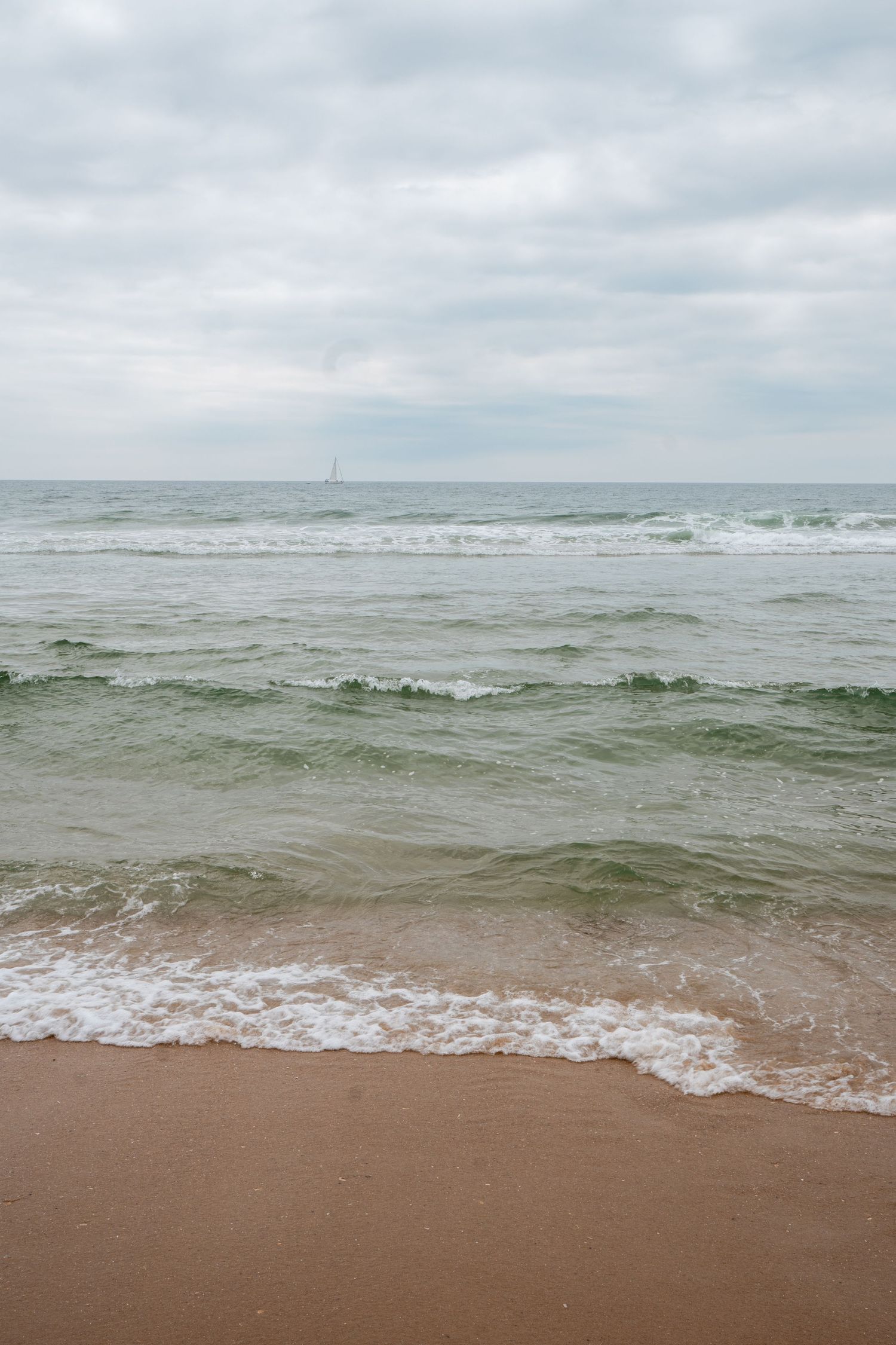 Calm ocean waves washing onto a sandy beach under an overcast sky, with a distant sailboat on the horizon.