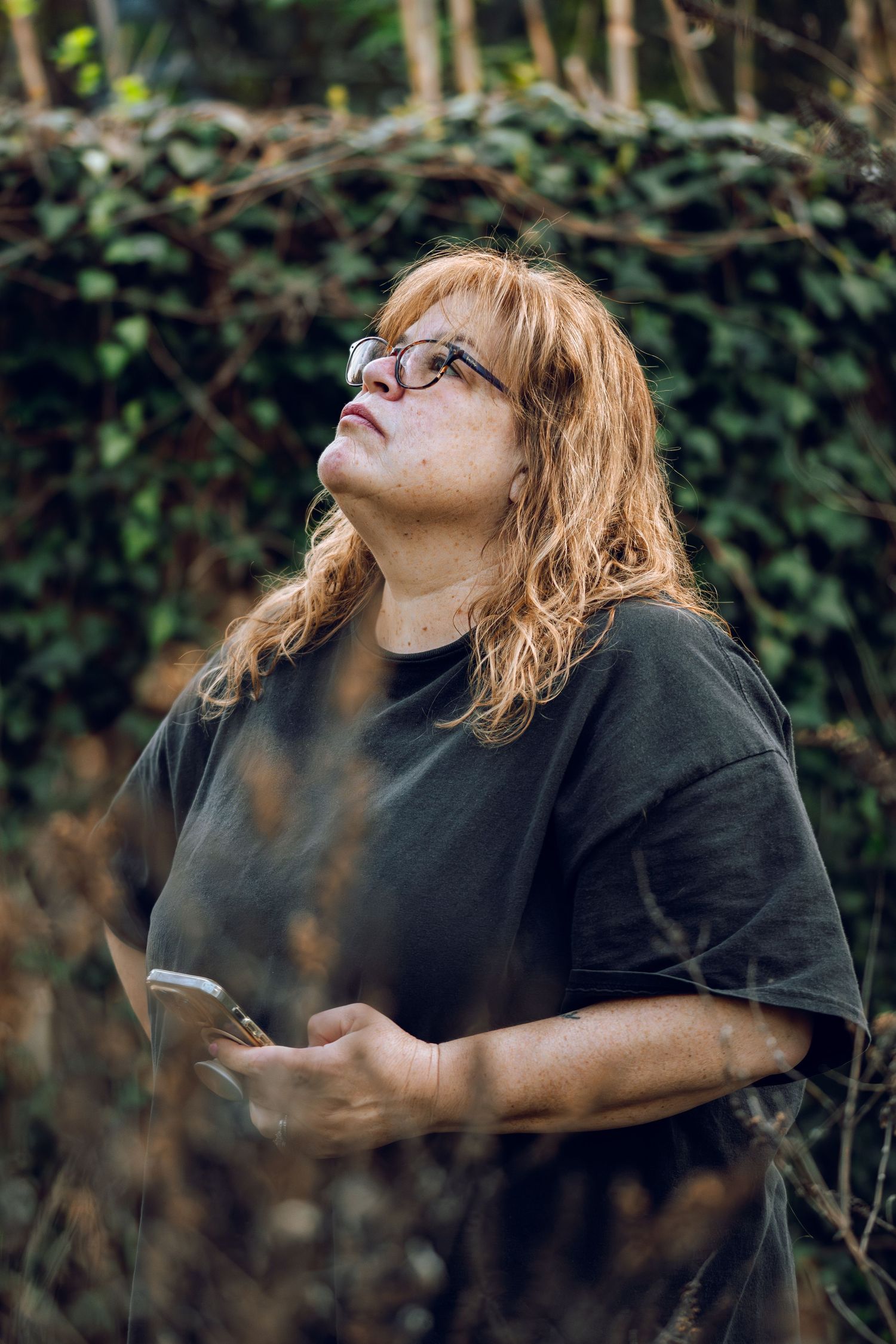 A red-haired woman wearing glasses and a black t-shirt looks upward while standing in front of green ivy.