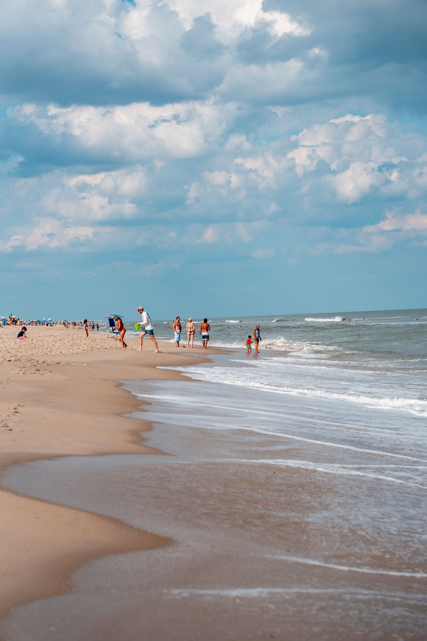 Beachgoers walking and playing along a wide sandy ocean shoreline under dramatic cloudy blue sky.
