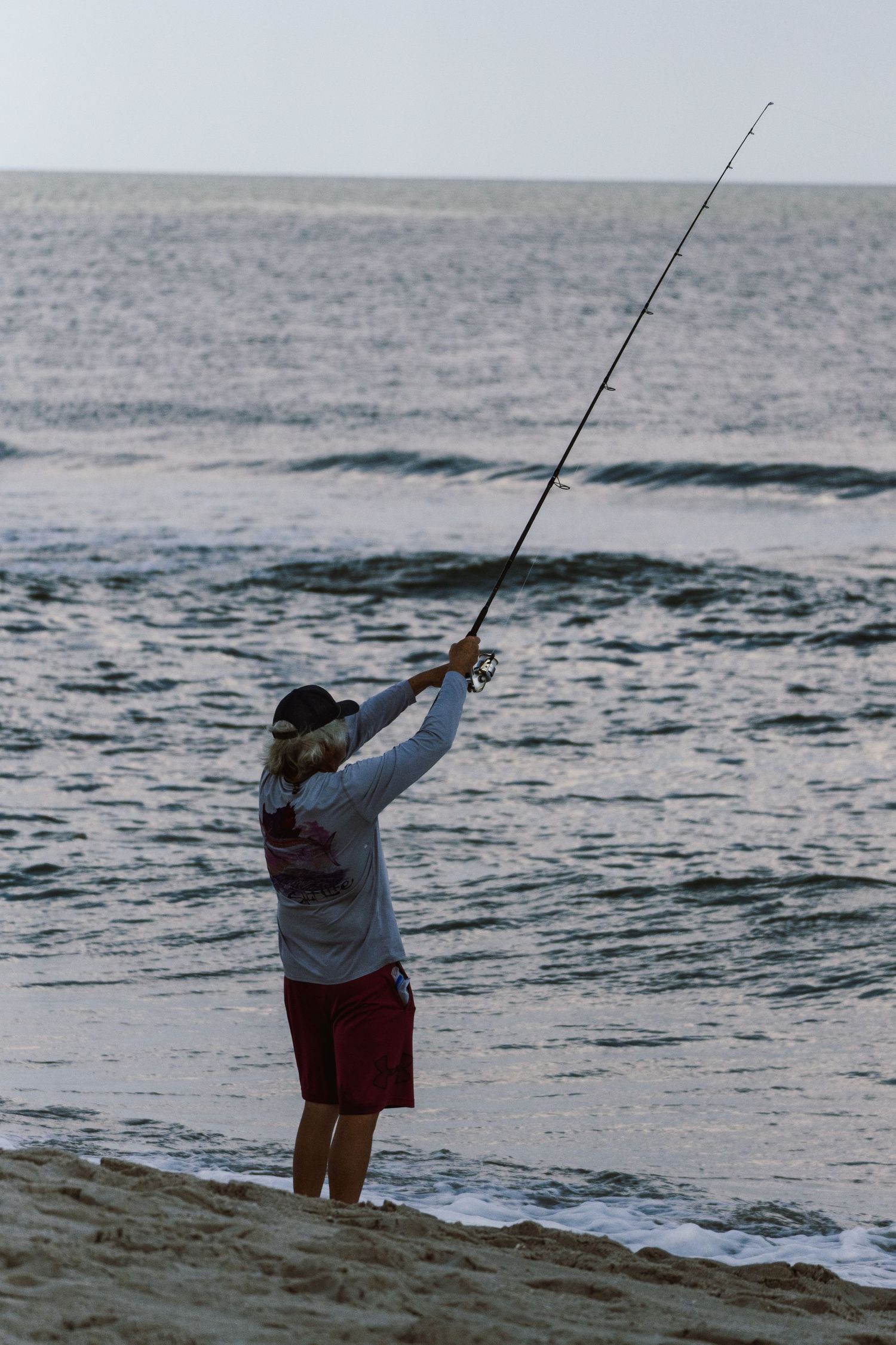 Person surf fishing on sandy beach at dusk, casting rod into ocean waves.