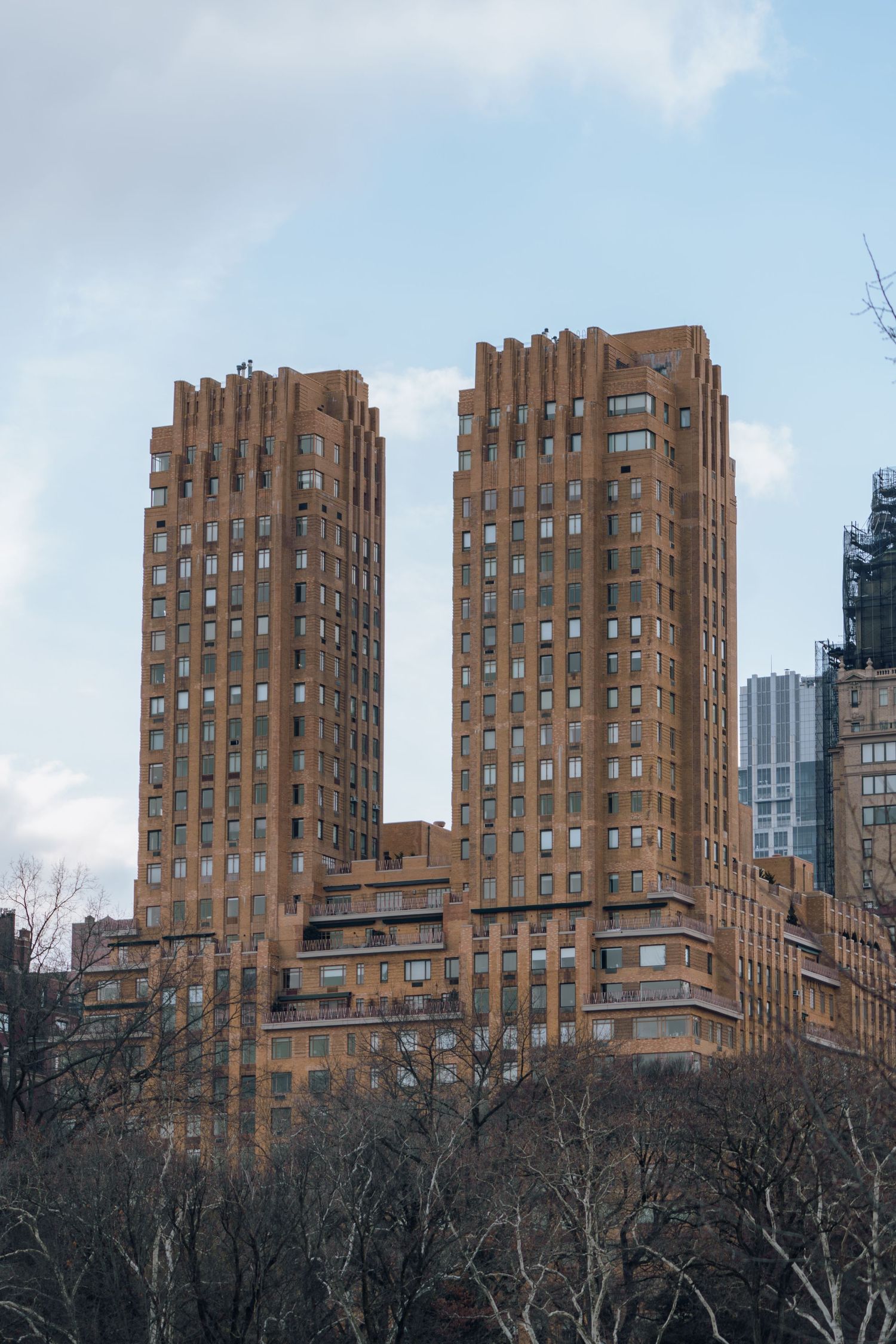 Twin brick residential towers with stepped facades overlook bare winter trees in Central Park, New York City.
