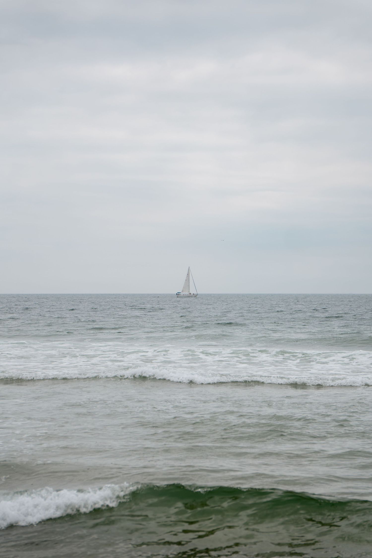 A lone sailboat glides across calm ocean waves under an overcast sky, viewed from a sandy beach shoreline.