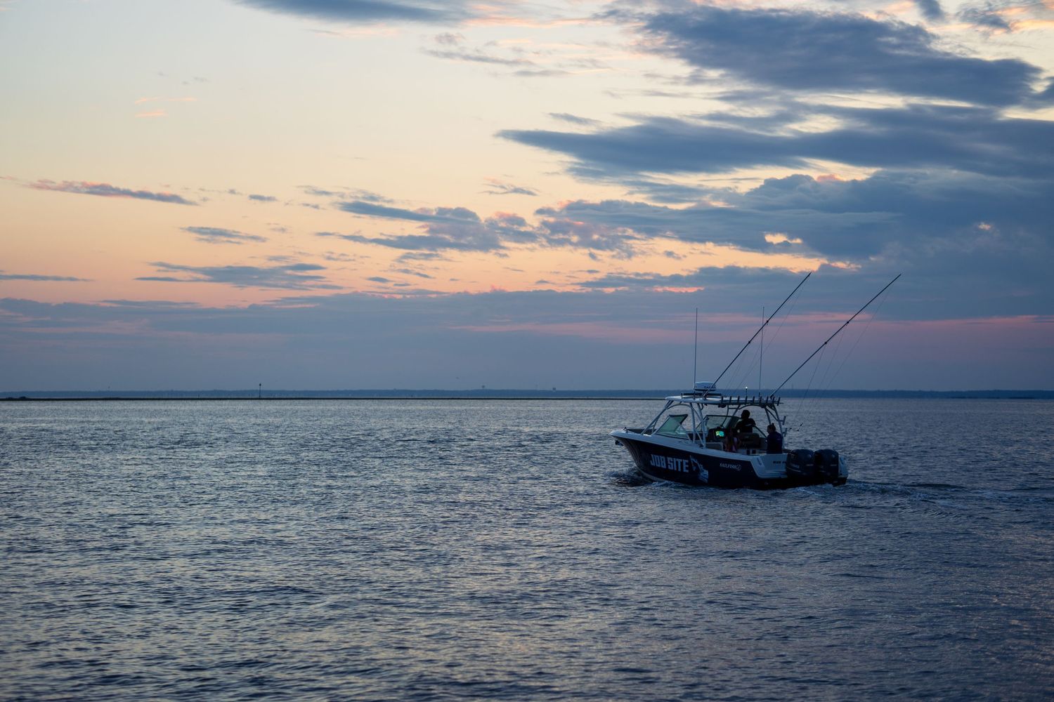 A fishing boat with tall rods silhouetted against a colorful sunset sky on calm ocean waters.