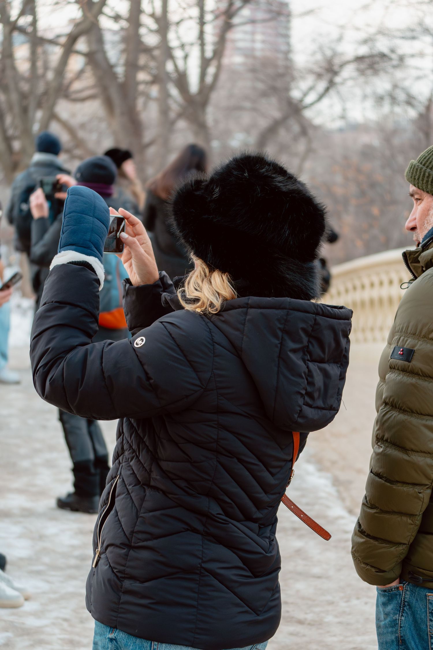 A woman in a black fur hat and puffer jacket photographs winter scenery in a crowded snowy park.