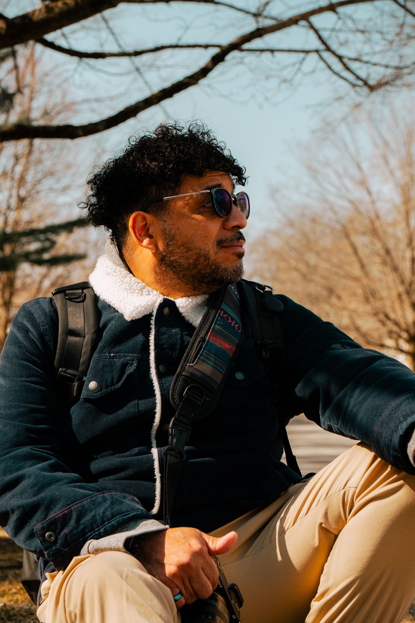 Curly-haired man in sunglasses and denim jacket with backpack sitting outdoors under bare winter trees.