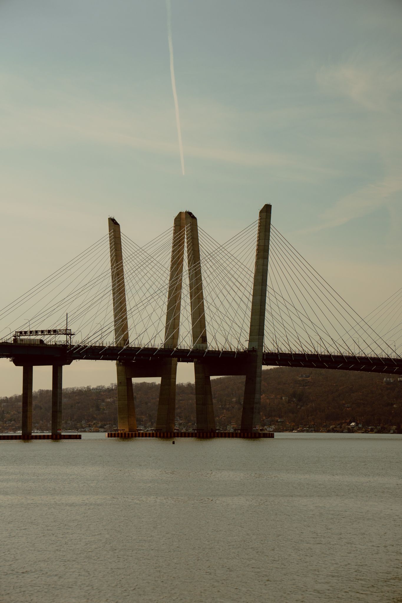 Cable-stayed bridge with tall pylons over river with airplane contrail in blue sky.