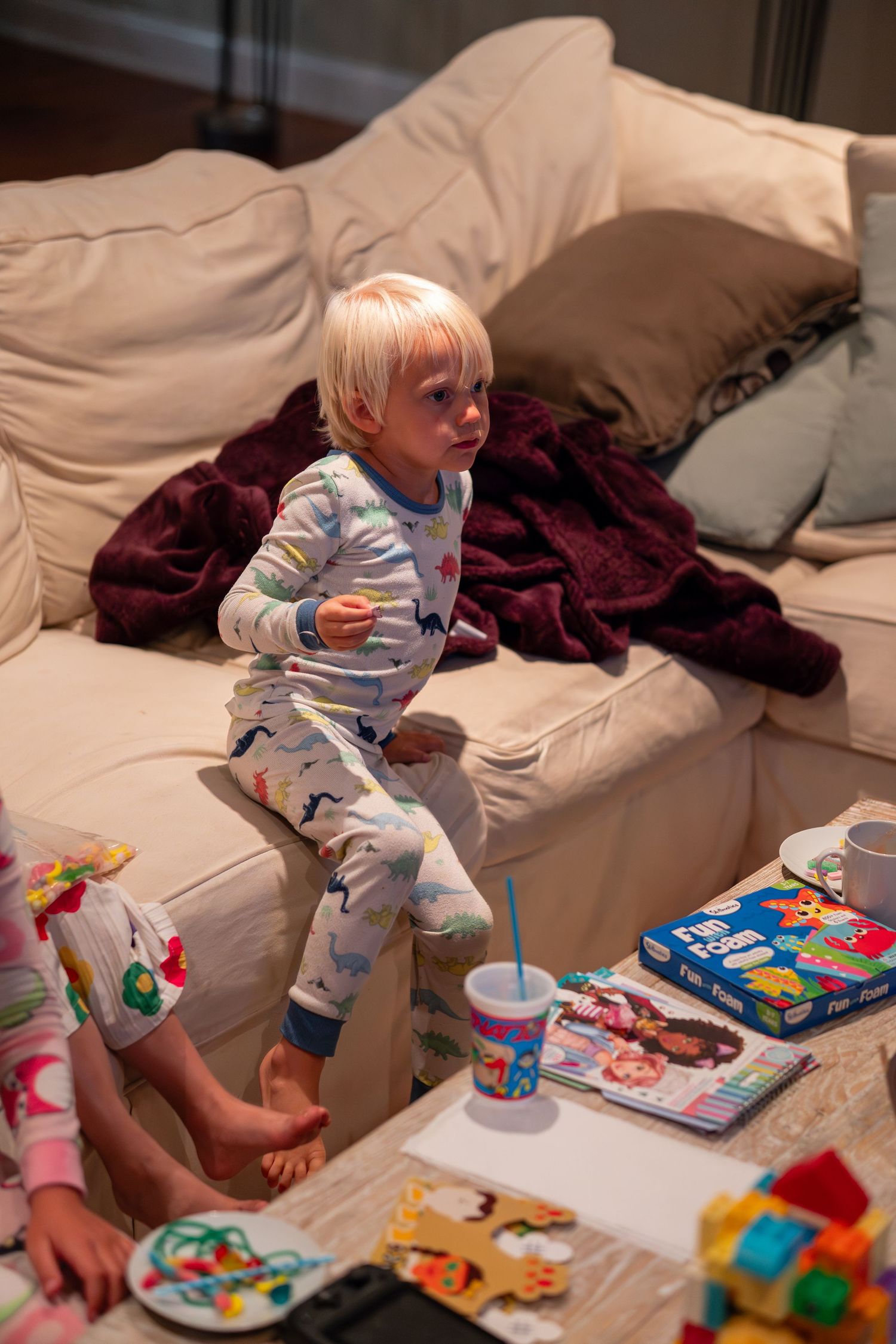 A blonde toddler in dinosaur pajamas sitting on a white couch with toys nearby.