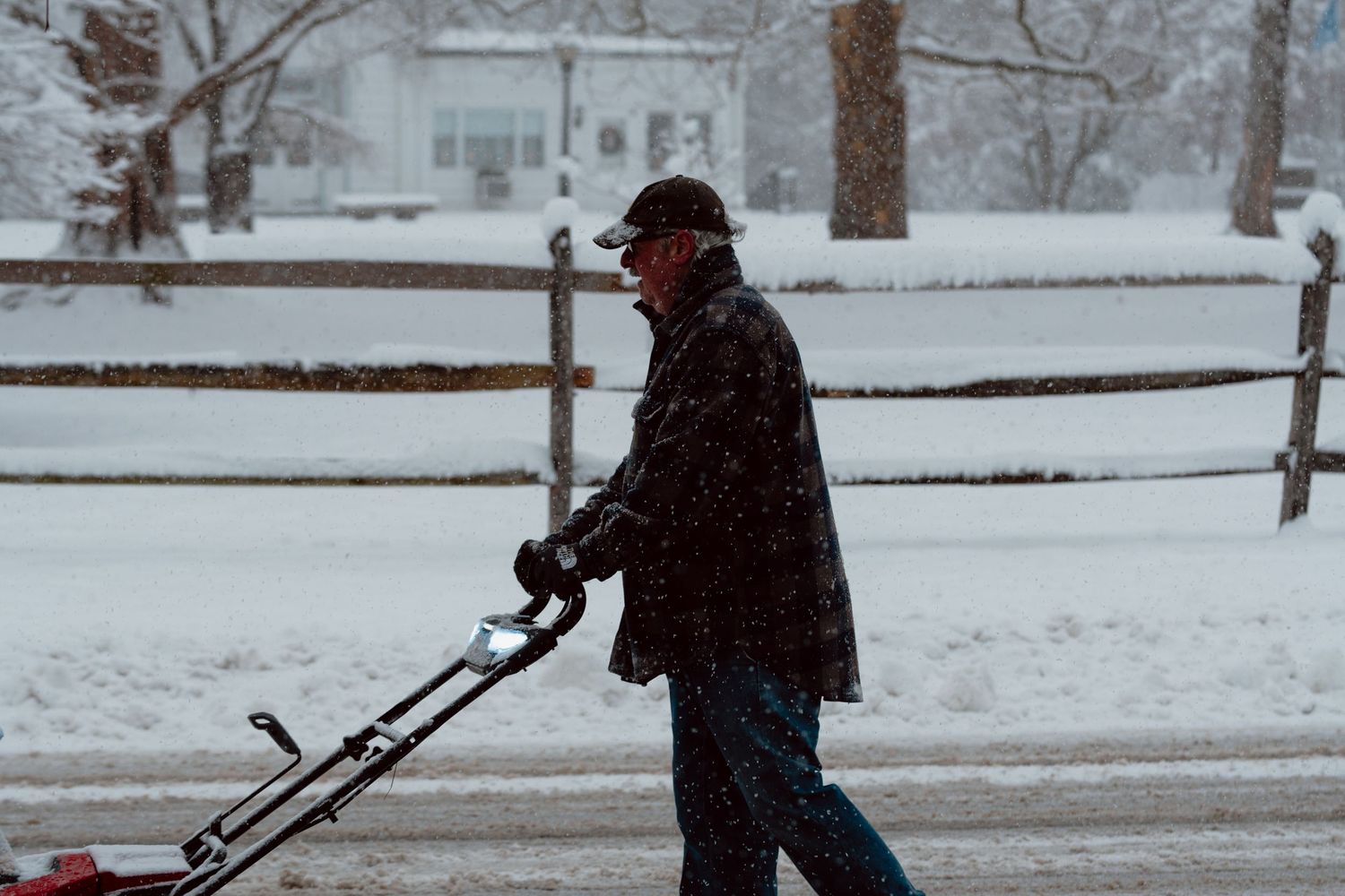 An elderly man using a red snowblower to clear a driveway during a heavy snowstorm.