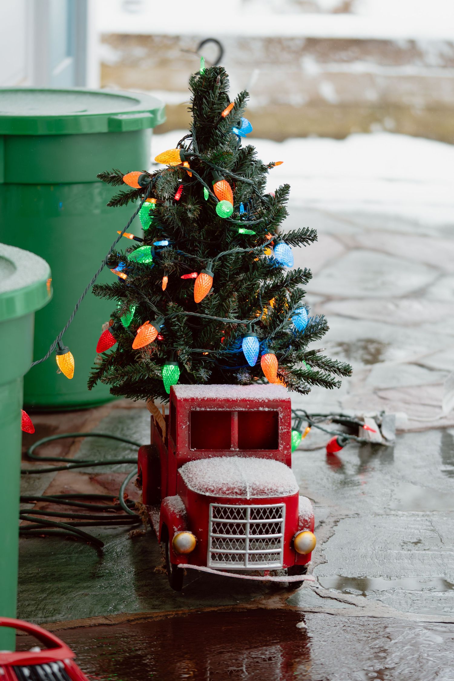 A decorative red toy truck carries a small Christmas tree adorned with colorful lights on a wet surface.