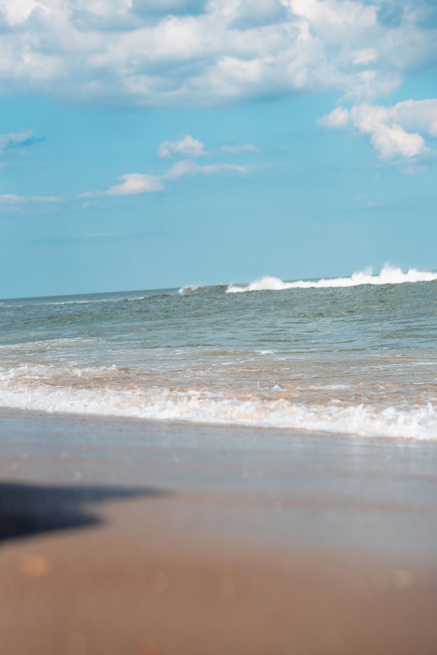 Ocean waves breaking on a sandy beach shore with fluffy white clouds in a bright blue sky.
